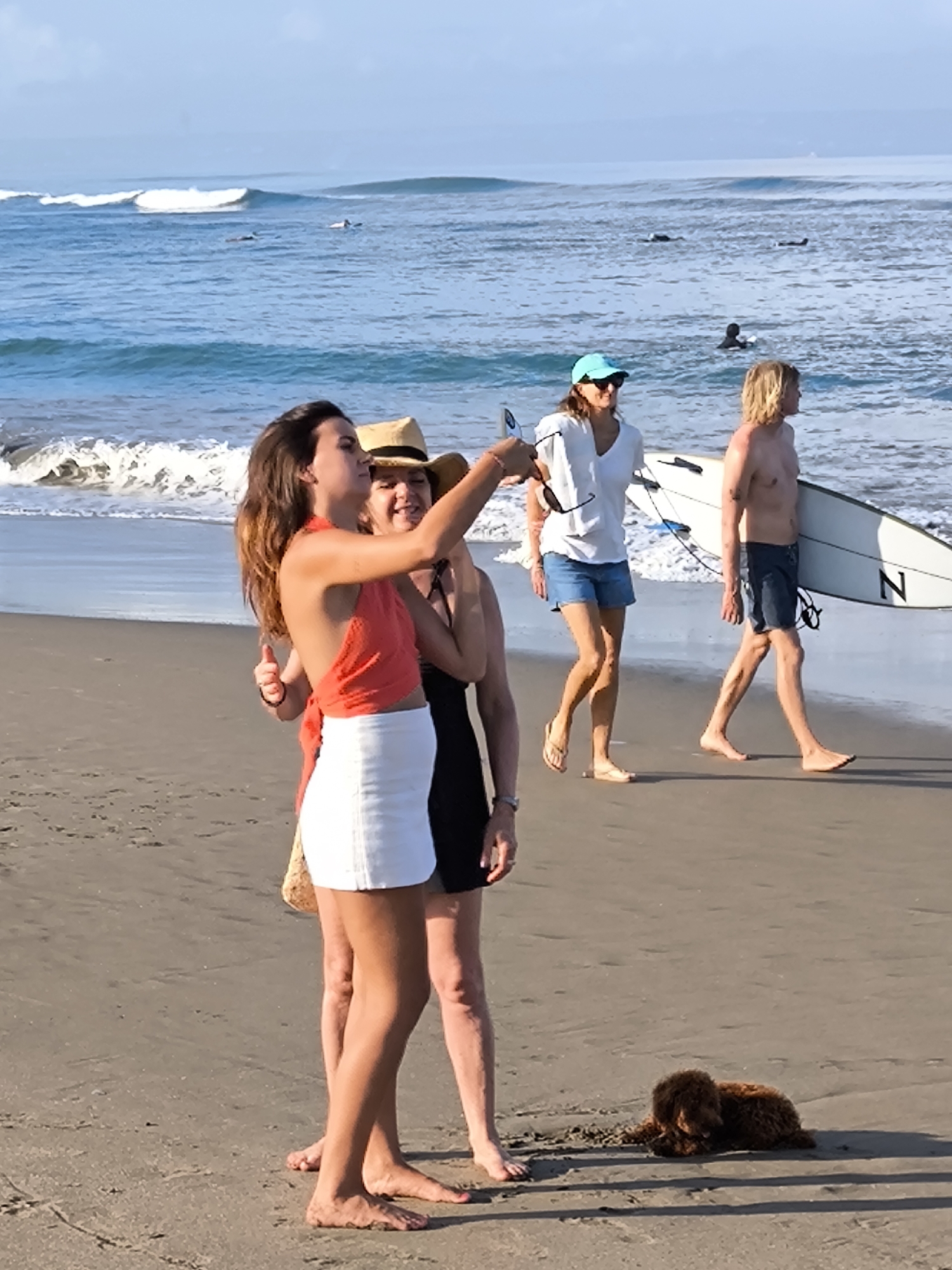 The photograph was taken on a beach in North Kuta, Indonesia. In the foreground, a woman in a red top and white skirt is taking a selfie with two other women, one in a hat and the other in a black dress. In the background, a woman in a blue hat, white t-shirt, and denim shorts is walking towards the right while holding a pair of sunglasses. Next to her, a shirtless man with a surfboard is also walking towards the right. In the far distance, there are surfers in the ocean. A small brown dog is lying on the sand near the group of women taking a selfie. The ocean waves are visible in the background.