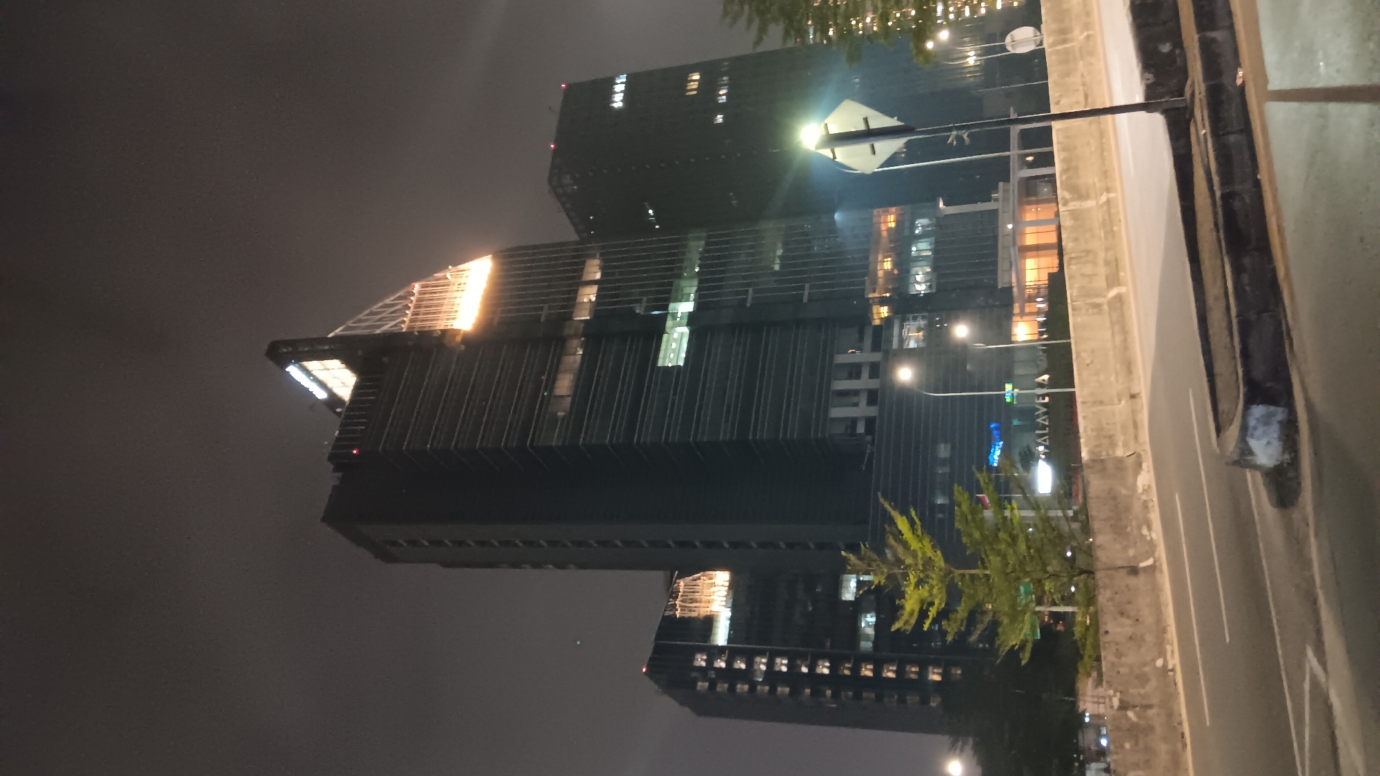 This image captures a nighttime view of the Alamanda Tower in South Jakarta, Indonesia. The focus is on the illuminated skyscraper, which is the central subject. The tower features a sleek, modern design with numerous windows reflecting the city lights. A distinctive angled top section of the tower is brightly lit, creating a striking contrast against the dark sky.
To the left of the main tower, another, slightly shorter building is visible, also illuminated with multiple lit windows. In the foreground, a multi-lane road is seen, with the road surface appearing wet, possibly from recent rain. A concrete barrier separates the road from the area in front of the buildings, where a young tree with bright green foliage stands out against the darker background. Streetlights are present, casting a warm glow on the scene. The overall atmosphere is urban and sophisticated, typical of a commercial district at night. No people are visible in the frame.