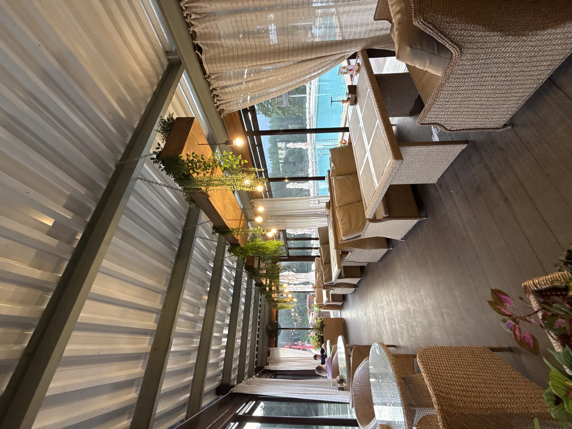 The image depicts an outdoor seating area of a cafe or restaurant with a view of a swimming pool. The seating area is covered by a corrugated metal roof supported by a metal frame. Wicker furniture, including tables and chairs with beige cushions, is arranged throughout the space. Several hanging plants adorn the ceiling. Large white curtains are drawn along the windows, partially obscuring the view.
Through the windows, a bright blue swimming pool is visible, with lounge chairs and umbrellas around it. In the background, lush green trees and hills can be seen, suggesting a natural or park-like setting. A couple is seated at a table in the foreground, facing away from the camera. Another person is visible further in the background, also seated. The overall atmosphere appears relaxed and resort-like. The location is identified as Barangol, Russia.