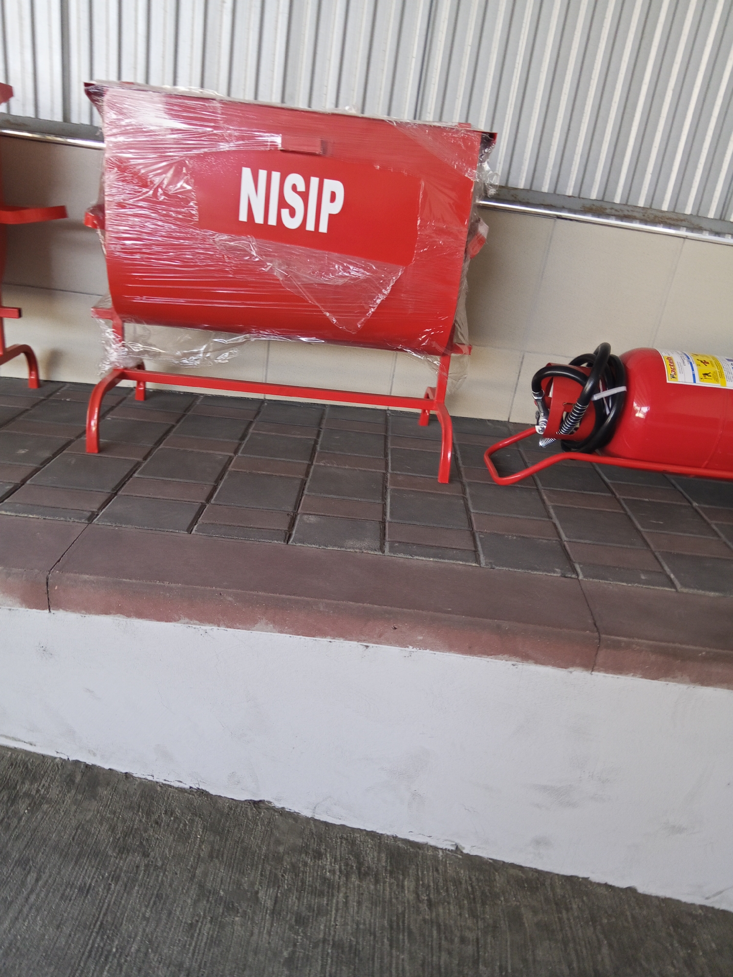 A red fire extinguisher, on its side, is positioned on a paved surface in front of a red metal stand. The fire extinguisher has a black hose and nozzle attached, and a label with yellow and white text is visible on its cylinder. To the left of the stand, a large red container, wrapped in clear plastic, is also on the stand. The word "NISIP" is printed in white on the front of this container. The background consists of a corrugated metal wall. The scene appears to be outdoors, possibly in a public area or storage facility, with a concrete sidewalk or base in the foreground. The location is Ceadîr-Lunga, Moldova.