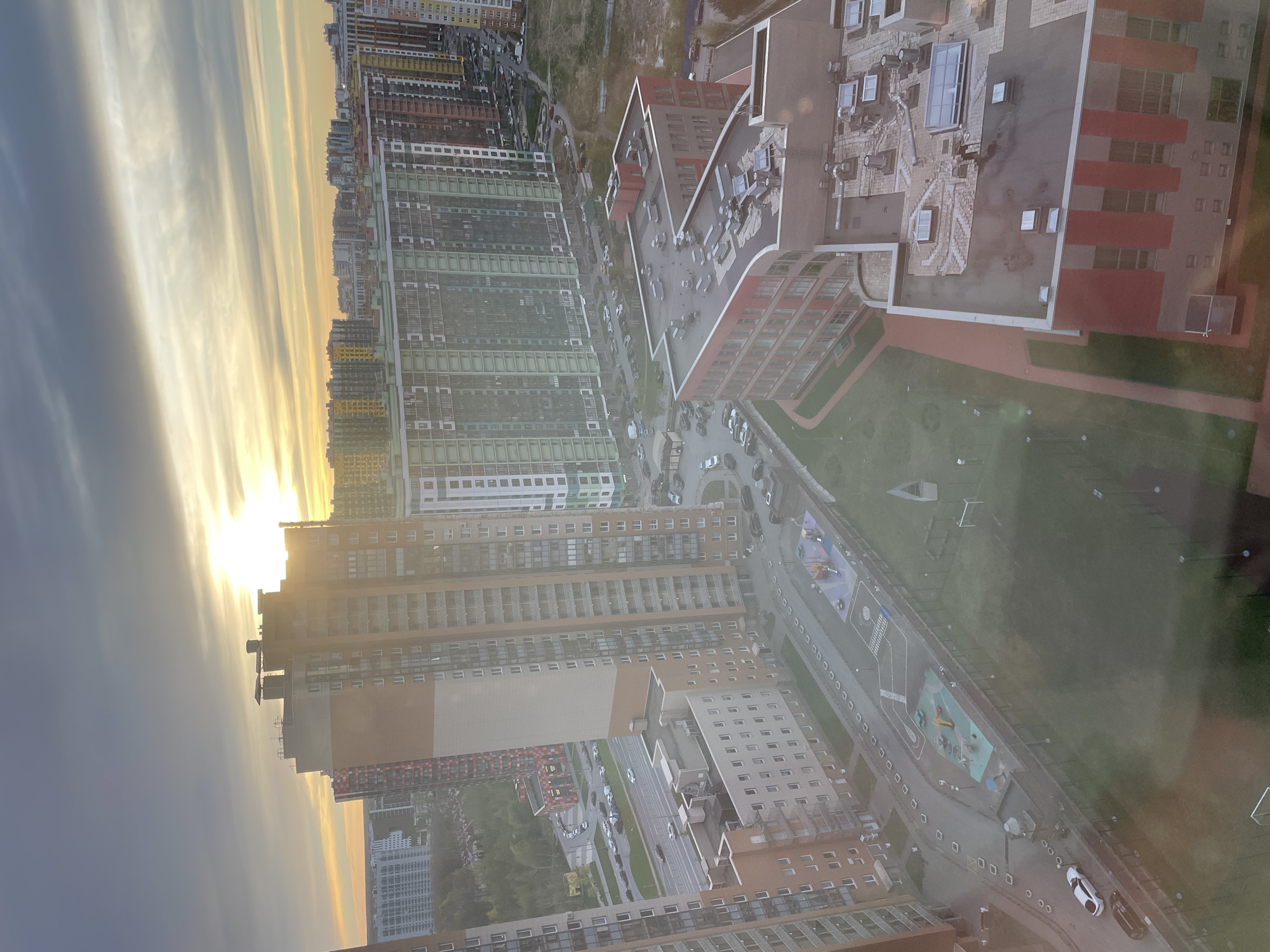 The image shows a high-angle view of a cityscape at sunset, likely taken from an elevated position within a building. The location is Saint Petersburg, Russia, identifiable by the architectural style and urban planning.
Buildings: Numerous high-rise residential buildings, many of which are multi-colored, dominate the scene. The buildings have uniform window patterns, suggesting modern apartment complexes. Several have red and gray facades and a cluster has green facades, with a variety of architectural styles.
Sky: The sky displays a sunset with hues of yellow and orange, with streaks of darker gray clouds.
Roads and infrastructure: A road is visible with cars parked along its side. There is a children's playground area with colorful installations.
Environment: A green area with a manicured lawn is present, indicating a planned urban space, possibly a park or courtyard. The overall scene conveys a modern urban residential area.