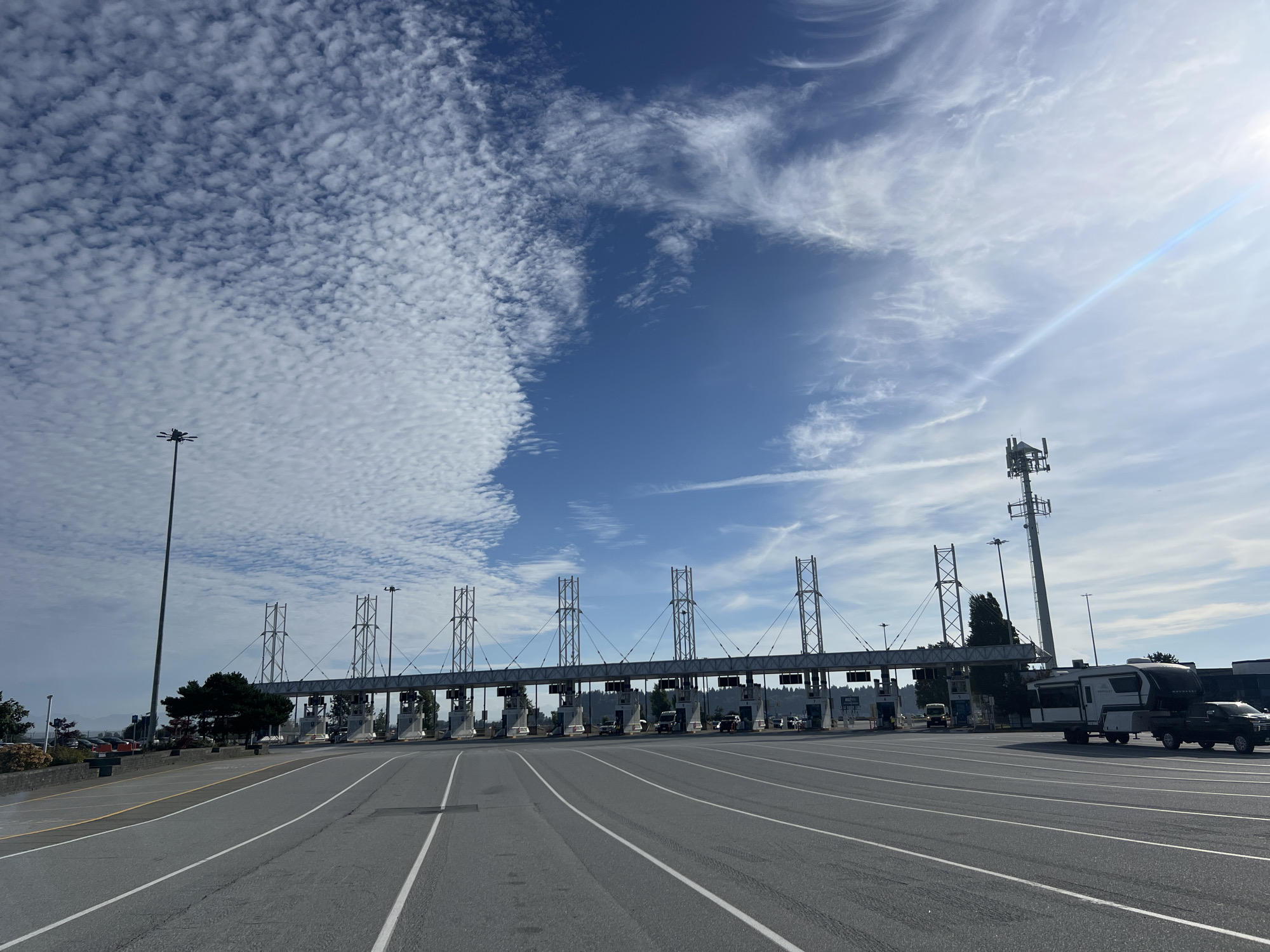 The image captures a view of the Tsawwassen ferry terminal in Delta, Canada, under a bright, partly cloudy sky. The foreground shows a wide, multi-lane asphalt road with white lane markings, leading towards a series of toll booths. The toll plaza features concrete pillars supporting overhead structures with multiple lanes. Several vehicles, including cars, SUVs, and a large RV being towed by a pickup truck, are visible, either waiting at the toll booths or driving through. In the background, a treeline and distant hills are visible. A tall communications tower with antennas stands to the right of the toll plaza. The sky is characterized by a mix of deep blue and scattered white, fluffy clouds, with the sun creating a lens flare on the right side of the frame. The scene is one of a busy transportation hub on a clear day.