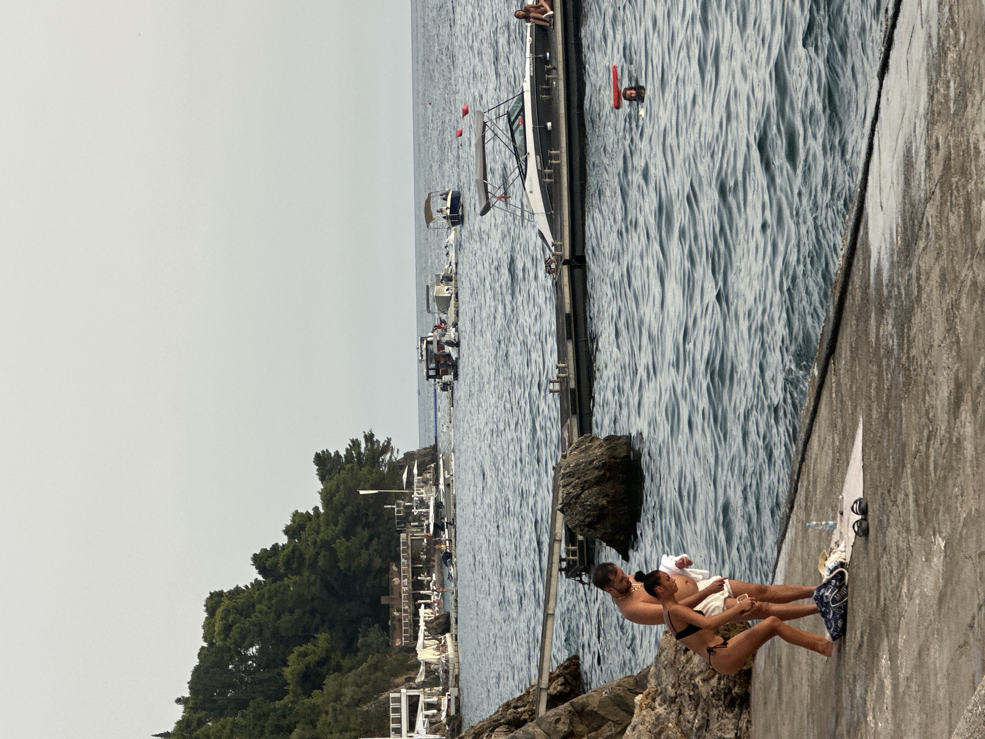 A couple is standing on a concrete walkway next to a rocky shore and the sea. The man is shirtless and wearing white shorts, while the woman is wearing a black bikini. The woman is bending down, looking at her phone. A white towel and a pair of black flip-flops are on the ground next to them. A water bottle is also on the ground. In the background, there is a dock with several boats and people, as well as buildings on a wooded hillside. The sea is calm with some small waves. The sky is overcast. This scene is likely at Mogren Beach in Budva, Montenegro, a popular tourist destination known for its rocky shores and clear blue waters.