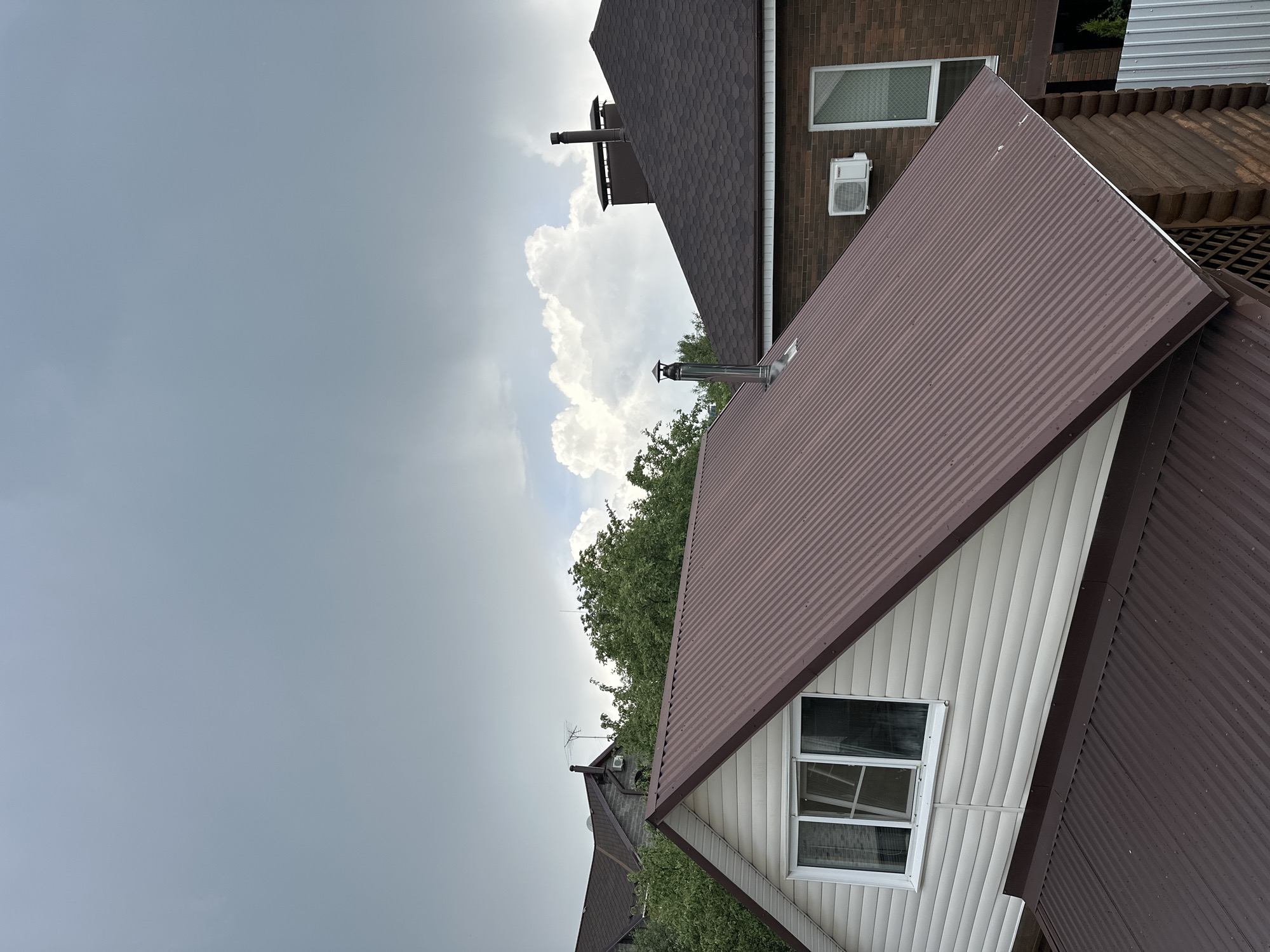 The image captures an exterior view of two residential houses, primarily focusing on their roofs and the sky above, set in Nizhny Novgorod, Russia. The perspective suggests a slightly elevated viewpoint, looking across rooftops towards the horizon.
In the foreground and to the right, a house features a brown corrugated metal roof, angled steeply. Its visible side wall is covered in white vertical siding, and a rectangular window with a white frame is centrally positioned on this wall. A metallic chimney or vent pipe protrudes from this roof.
Behind and to the left of the first house, another house is partially visible. It has a dark brown shingled roof with a distinct hexagonal pattern. Its wall is constructed of brown brick, and it also features a rectangular window with a white frame, accompanied by an outdoor air conditioning unit mounted directly below it. A darker, box-like chimney structure is visible on this house's roof.
Green tree foliage is scattered throughout the mid-ground, peeking out between the houses and below the cloud line. The sky above presents a contrast: the upper left portion is largely overcast with a uniform grey tone, while the central and right parts feature large, bright, white cumulus clouds against a clearer, lighter sky, indicating daytime.
There is no specific activity taking place; the image is a static architectural and landscape shot. No visible text is present in the scene. The mixed sky conditions suggest a day with varying weather, possibly transitioning between sunny and cloudy.