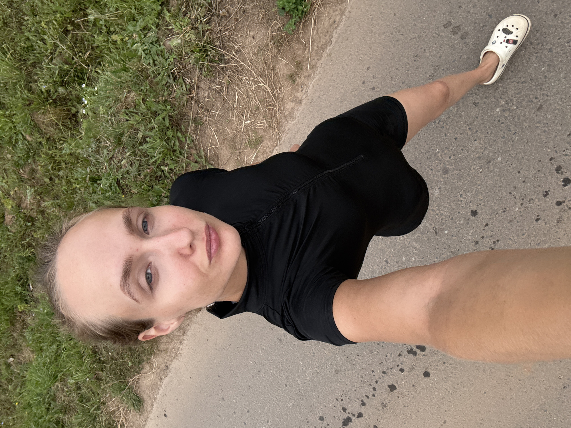 A young woman with fair skin and light brown hair pulled back is shown from a high-angle perspective, taking a selfie outdoors. She is wearing a black, short-sleeved, collared top with a zipper down the front and white clogs with cartoon designs. The woman is standing on a paved surface, possibly a road or pathway, with a grassy area on her right. The ground is uneven and shows signs of wear, with dark splotches. The grassy area is overgrown with various types of vegetation and some dry branches. The overall lighting suggests it is daytime, likely overcast or late afternoon, with soft shadows. The location is identified as Podshivalovo, Russia.
