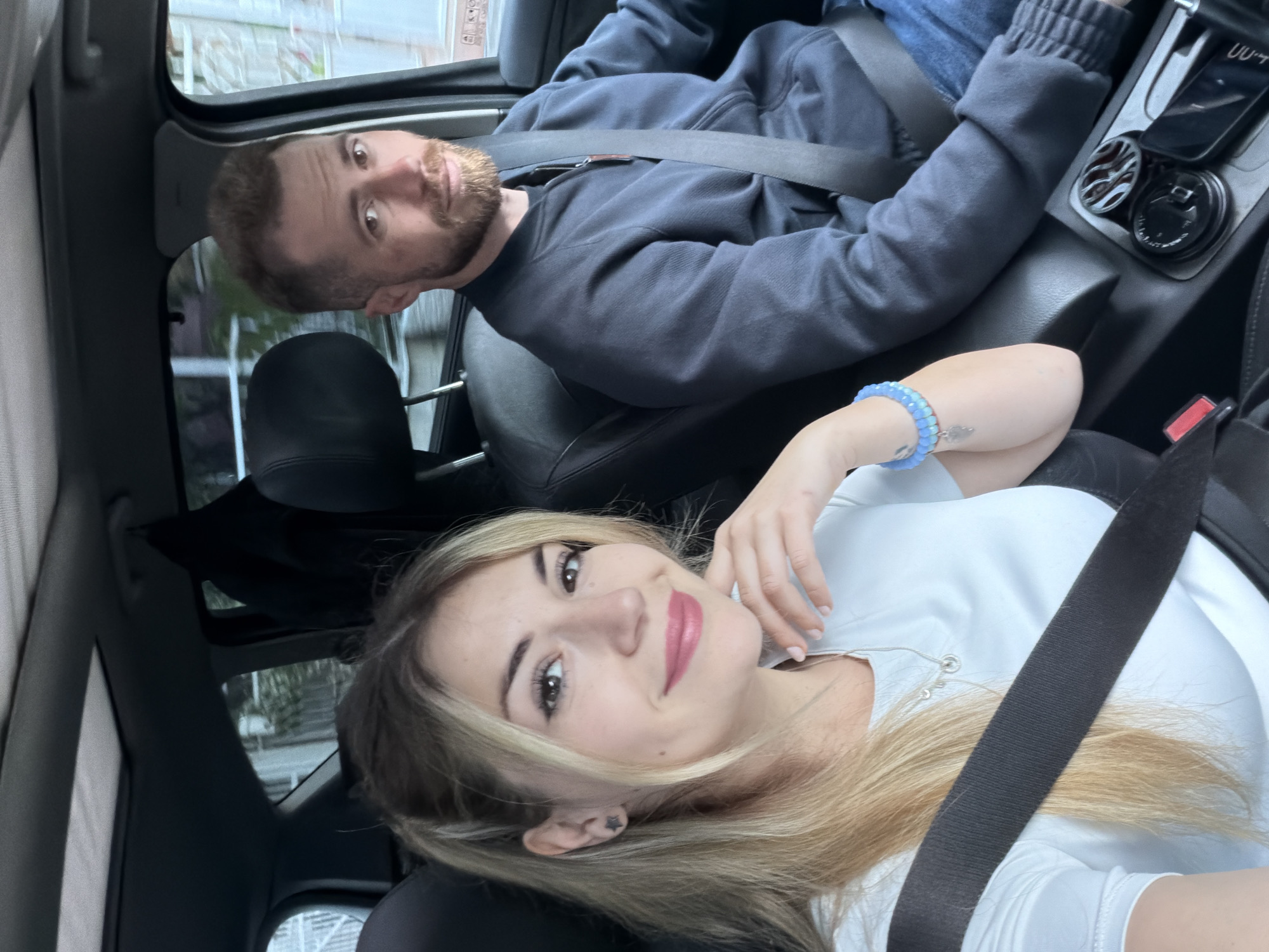 Inside a car, a blonde woman wearing a white shirt is in the passenger seat, smiling and facing the camera. She has a blue bracelet on her wrist and is wearing a seatbelt. A man with a beard is in the driver's seat, also wearing a seatbelt. The interior of the car is black. Outside the car windows, blurred buildings and trees suggest a city environment, consistent with the style seen in the city of Budva, Montenegro.