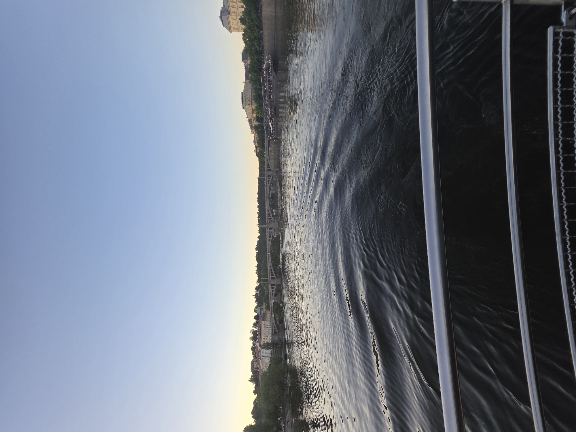 The image captures a river scene, likely from a boat or a waterfront location, in an unidentified city. A large body of water, possibly a river or a wide waterway, dominates the scene. The water surface shows ripples and waves, indicating movement or wind. The shoreline features buildings and structures, including a bridge extending across the water. The sky is clear and bright, suggesting either morning or afternoon lighting conditions. In the foreground, parts of the boat's railing or structure are visible, providing perspective.