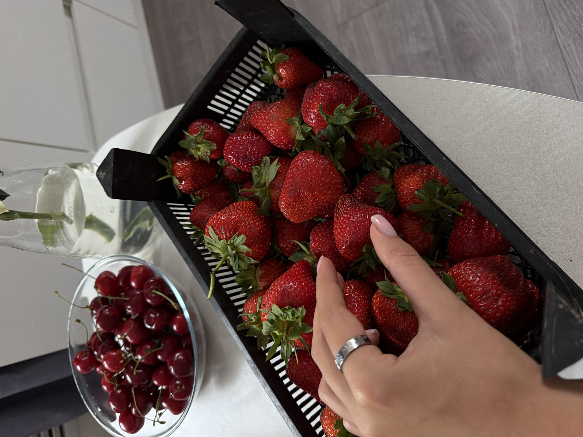 A high-angle shot displays a black rectangular basket filled with strawberries, beside a glass bowl of cherries, and a vase with greenery. The scene is set on a white table, against a background of a white cabinet and a grey wall. A human hand is partially visible, adorned with a ring, reaching into the basket of strawberries. The environment suggests an indoor setting, likely a kitchen. The photo was taken in Kazan, Russia.