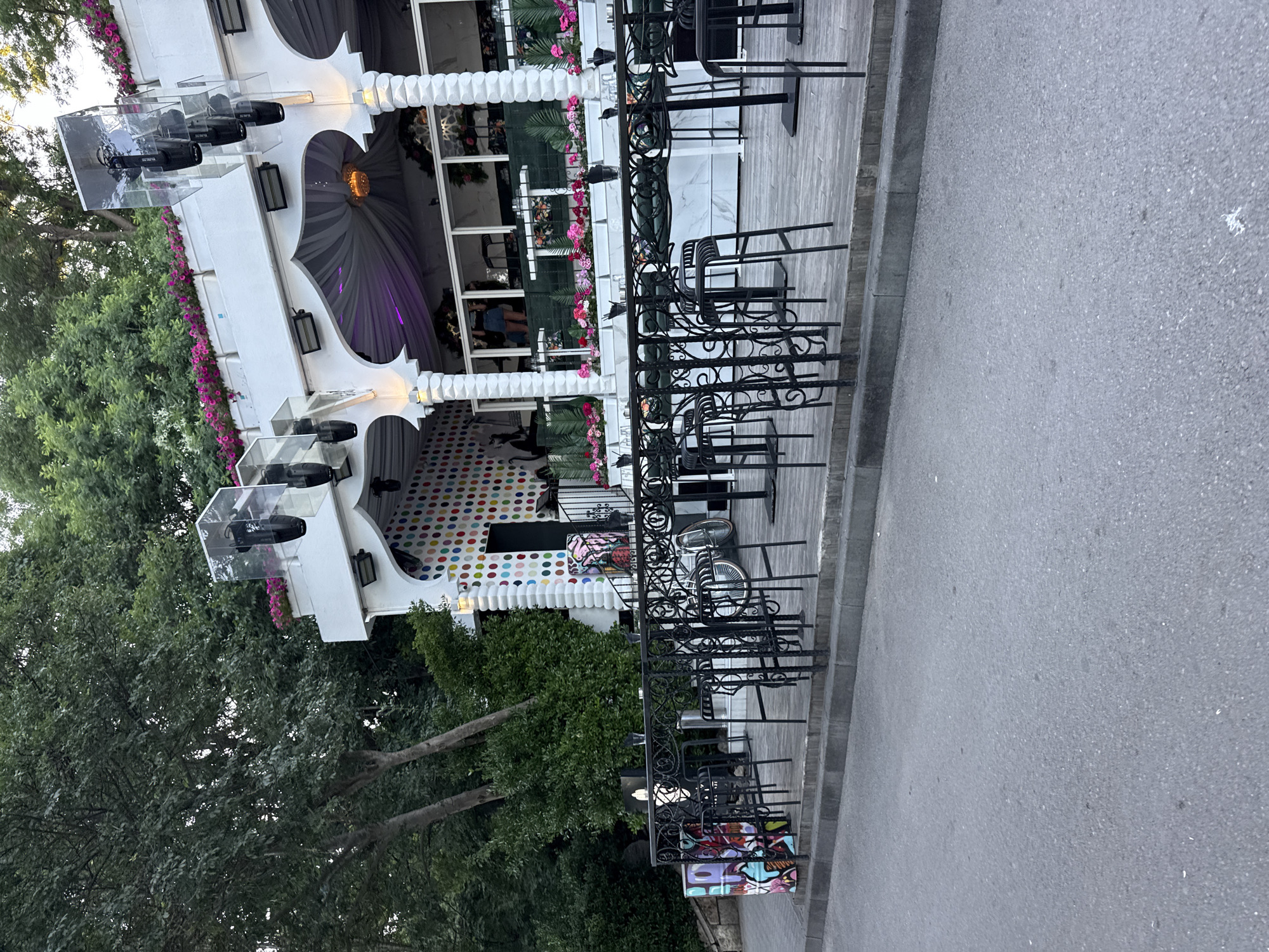 A wide shot captures an outdoor seating area of a modern establishment, possibly a cafe or bar, with a street in the foreground. The establishment features a white, multi-tiered structure adorned with floral arrangements and decorative pillars. The facade is decorated with a vibrant, polka-dot pattern. In front of the building, black metal tables and chairs are set up on a wooden deck. A street with a curb separates the viewer from the seating area. The background is filled with lush green trees. The lighting suggests it's daytime.