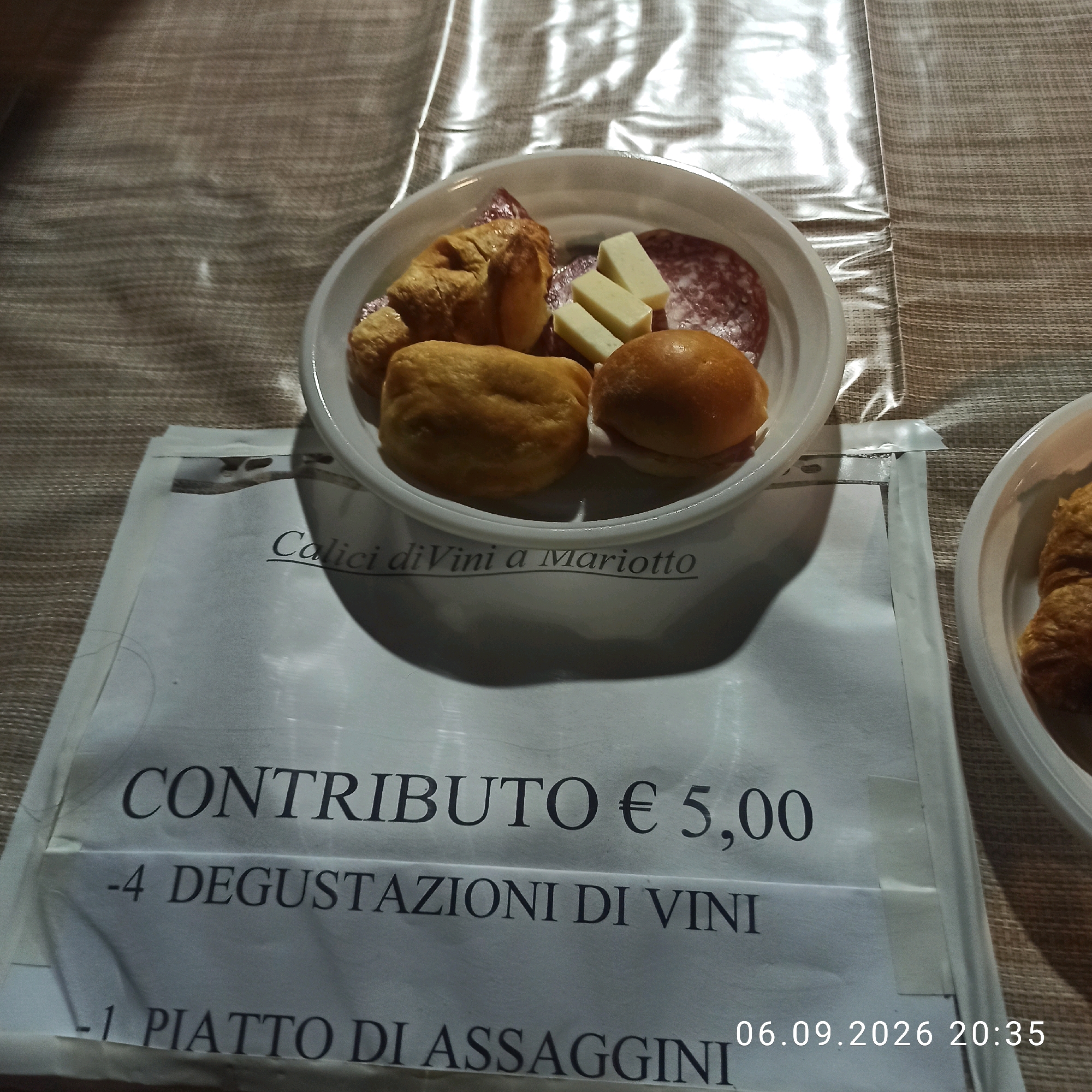 The image shows a white bowl filled with various food items, including croissants, bread rolls, cheese cubes, and salami. The bowl is placed on a textured brown surface, possibly a tablecloth. In front of the bowl, a white sheet of paper displays text. The text on the paper reads "Calici diVini a Mariotto", "CONTRIBUTO € 5,00", "-4 DEGUSTAZIONI DI VINI", and "-1 PIATTO DI ASSAGGINI". A date and time stamp, "06.09.2026 20:35", is visible in the bottom right corner of the paper. There are no people visible in the image. The lighting suggests it might be indoors or evening. Based on the text "Calici diVini a Mariotto," this likely refers to an event or establishment related to wine tasting in Bitonto, Italy, possibly a wine bar or an event organized by "Mariotto" within the city.