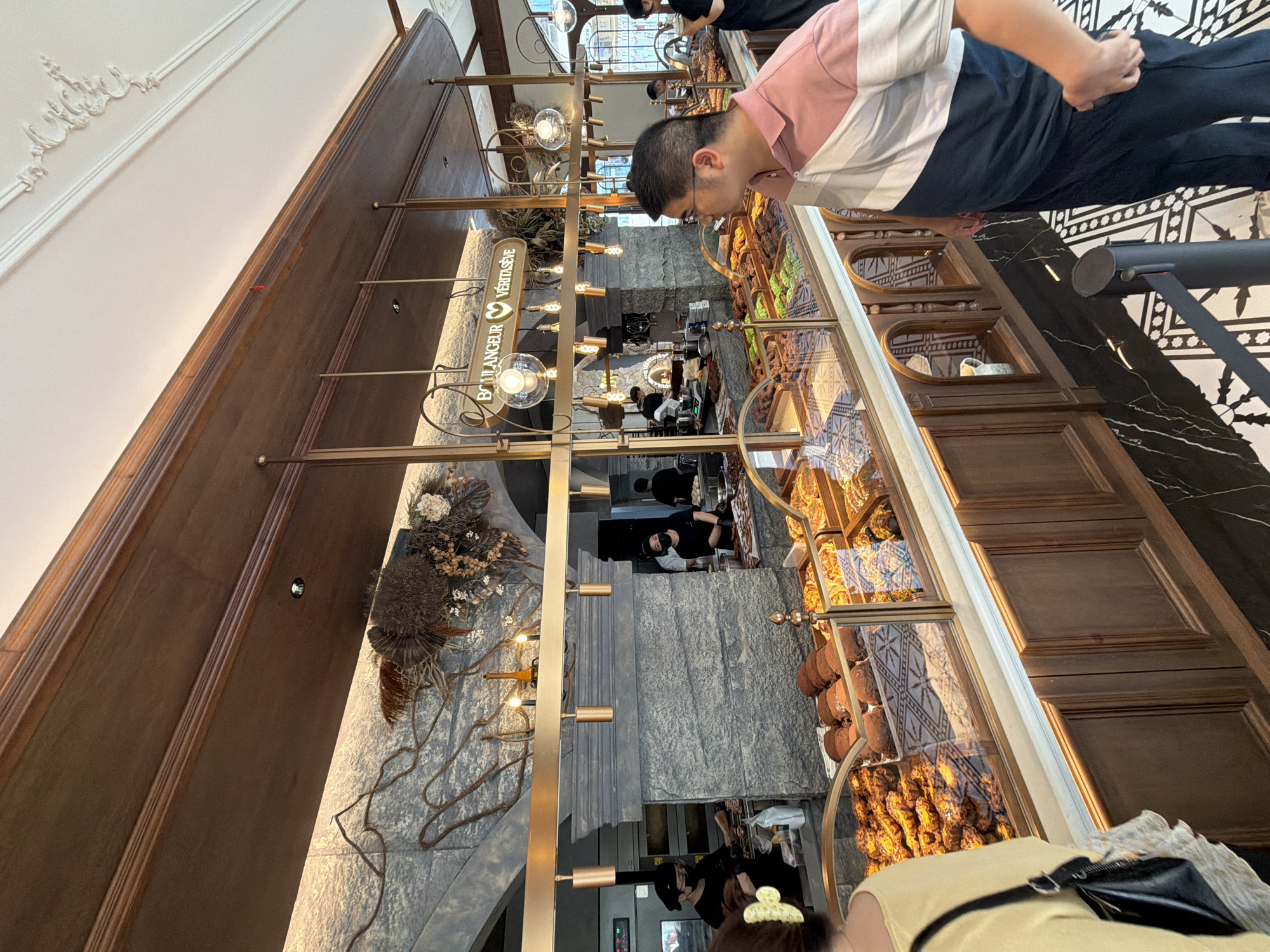 Here is a description of the image.
The image shows the interior of a bakery or pastry shop located in the Chongqing High-tech Zone, China. A man wearing a pink and white striped shirt and dark pants is leaning over a display case filled with various baked goods. The display case has a glass front and is adorned with intricate designs. Behind the display case, there are employees, some wearing masks, working in the kitchen area. A second person is partially visible in the foreground, wearing a yellow shirt and carrying a black purse. The shop has a modern and elegant design, with marble floors and walls, and ornate lighting fixtures. The bakery's name, "Boulangeur Veritaseve," is displayed on a sign.