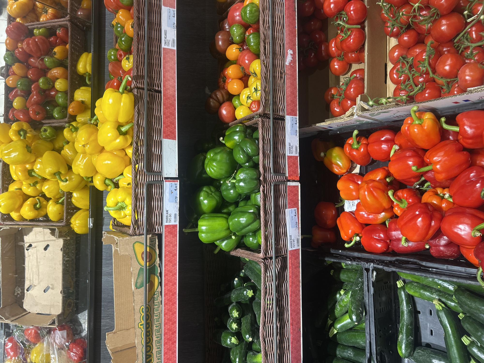 The image captures a vibrant fresh produce section, likely within a grocery store in Noisy-le-Grand, France. The scene features abundant displays of colorful vegetables neatly arranged in woven baskets, cardboard boxes, and black plastic crates.
The primary subjects are various fresh vegetables. Bright yellow bell peppers are prominent on the left, some filling woven baskets and others overflowing from a cardboard box. Above them, a diverse collection of mixed tomatoes in shades of red, orange, yellow, and green are visible. Green bell peppers are showcased in baskets in the middle section, while below them, dark green zucchini are stacked in plastic crates. On the right, numerous red and orange bell peppers are displayed alongside clusters of red tomatoes on the vine.
No people are visible, focusing the attention entirely on the produce. The setting is clearly a retail environment, with clear pricing and origin information for the goods.
Notable details include the organized presentation by color and type, the fresh appearance of the vegetables, and the distinct containers. An empty cardboard box with an avocado graphic is also visible. The lighting is bright and even, suggesting an indoor setting.
Visible text from the price labels includes:
- "POIVRON JAUNE PAYS BAS Le kg 4,99€" (Yellow bell pepper from the Netherlands, 4.99€ per kilogram).
- "TOMATE MELANGEES FRANCE Le kg 6.50€" (Mixed tomatoes from France, 6.50€ per kilogram).
- "POIVRON ROUGE PAYS BAS Le kg 4,99€" (Red bell pepper from the Netherlands, 4.99€ per kilogram).