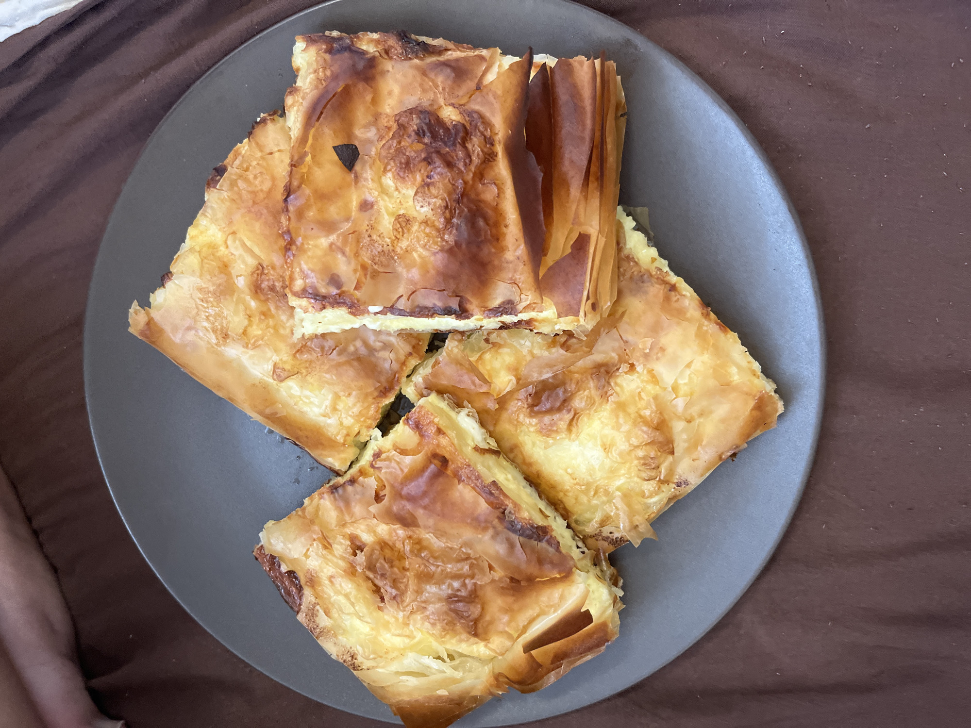 Four pieces of golden-brown phyllo pastry are arranged on a dark gray circular plate. The pastry is flaky with visible layers and a slightly charred, crispy texture in some areas. A creamy filling, likely cheese or custard, is visible in the cut sections. The plate rests on a dark brown, wrinkled fabric. The scene appears to be indoors, with soft lighting suggesting a home environment. There are no people visible in the image, and no specific landmarks or details indicate a location in Sofia, Bulgaria.