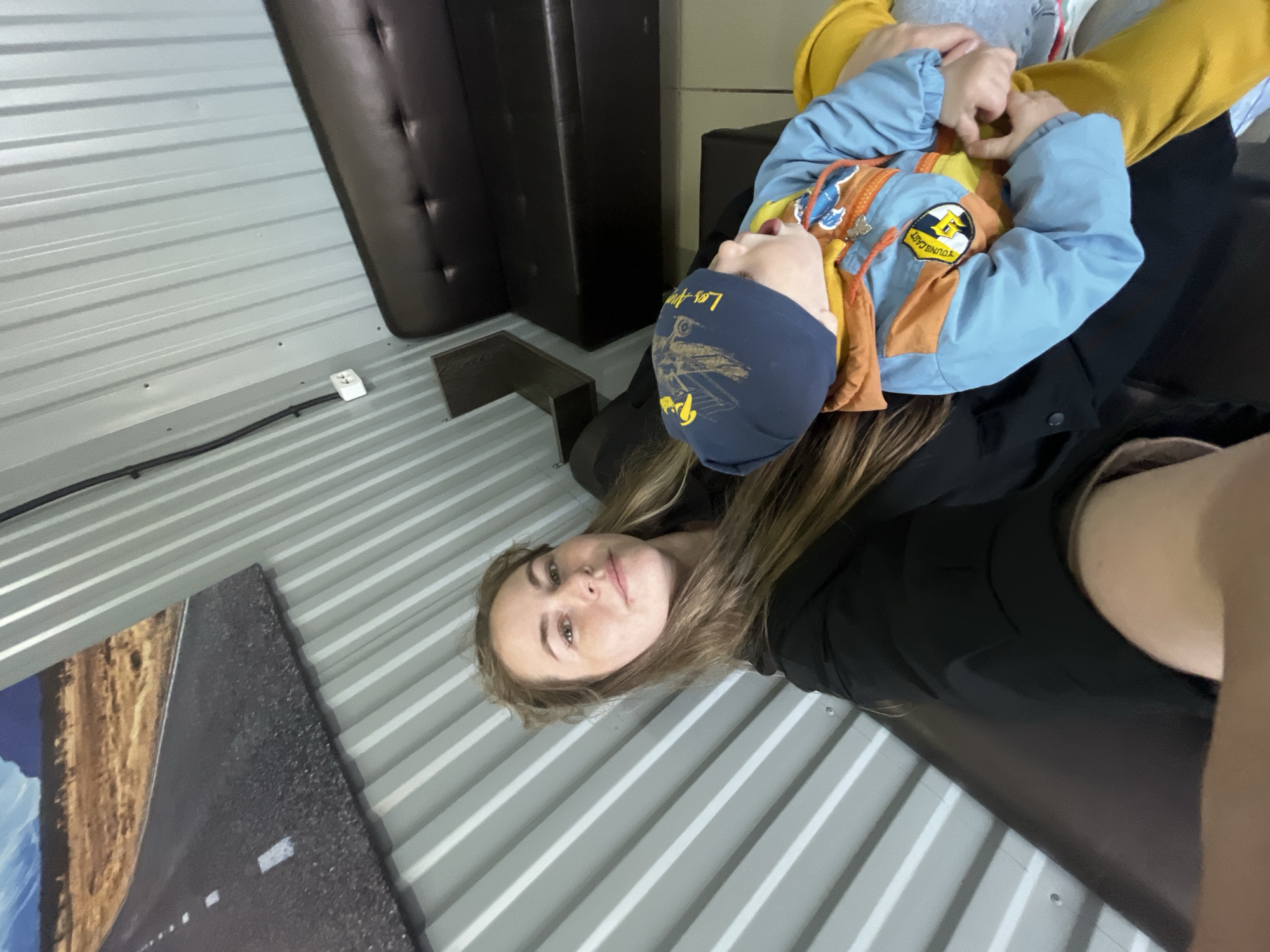 A woman and a child are inside a corrugated metal building. The woman has long, light brown hair and is wearing a black jacket. The child is wearing a blue hat, a blue and orange jacket, and yellow sleeves. The building has gray, corrugated metal walls and a dark brown leather couch. A framed photo of a road is visible on the left wall. There is an electrical outlet on the wall. The location is in the city of Chaikovsky, Russia.