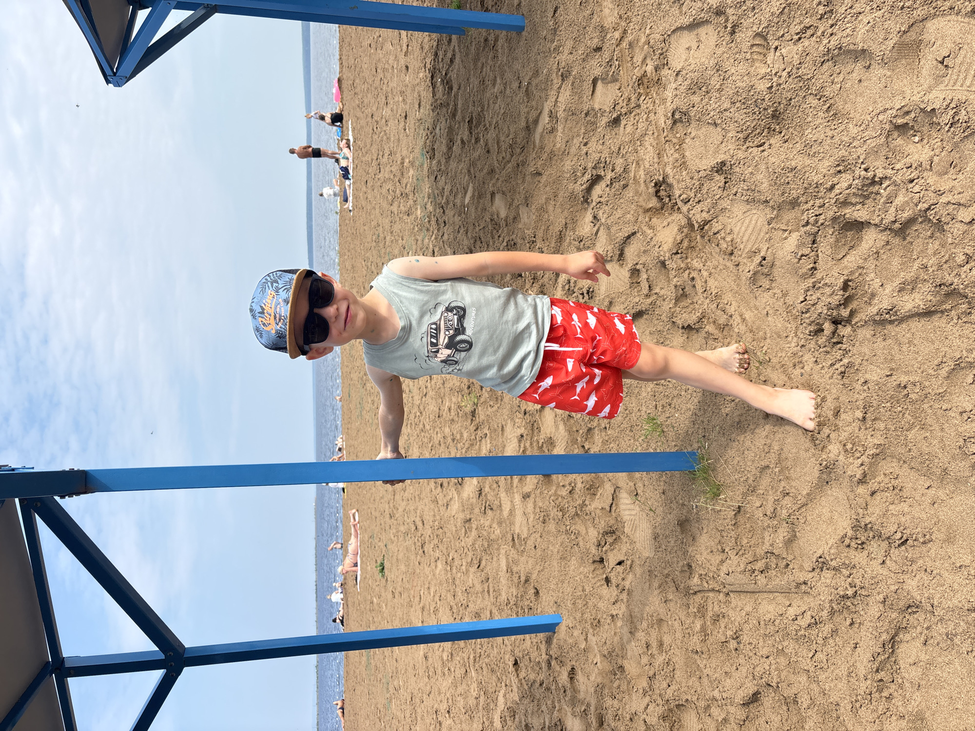 A young boy wearing a light grey tank top with a graphic of a jeep, red swim trunks with white shark prints, sunglasses, and a blue and brown patterned baseball cap stands on a sandy beach. He is leaning against a bright blue metal pole of an open-air shelter. The beach overlooks a body of water, likely a river or lake, with a distant shoreline and a few people visible in the background sunbathing and walking. The sky is partly cloudy. The scene is set in Chaikovsky, Russia, on the Kama River.