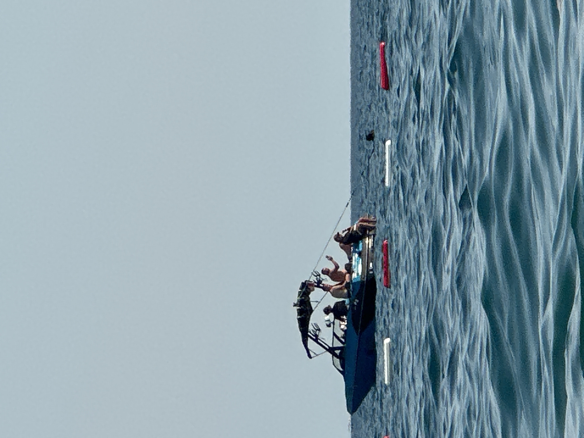 The image depicts a boat on the water near Boreti, Montenegro. The boat, a dark blue wakeboard boat, has a canopy and is occupied by five individuals. Four people are seated on the rear of the boat, and one person is seated in the front. The boat is surrounded by buoys, two white and two red, floating on the choppy blue water. The sky is clear and pale blue. The scene appears to be a recreational outing on the sea.
