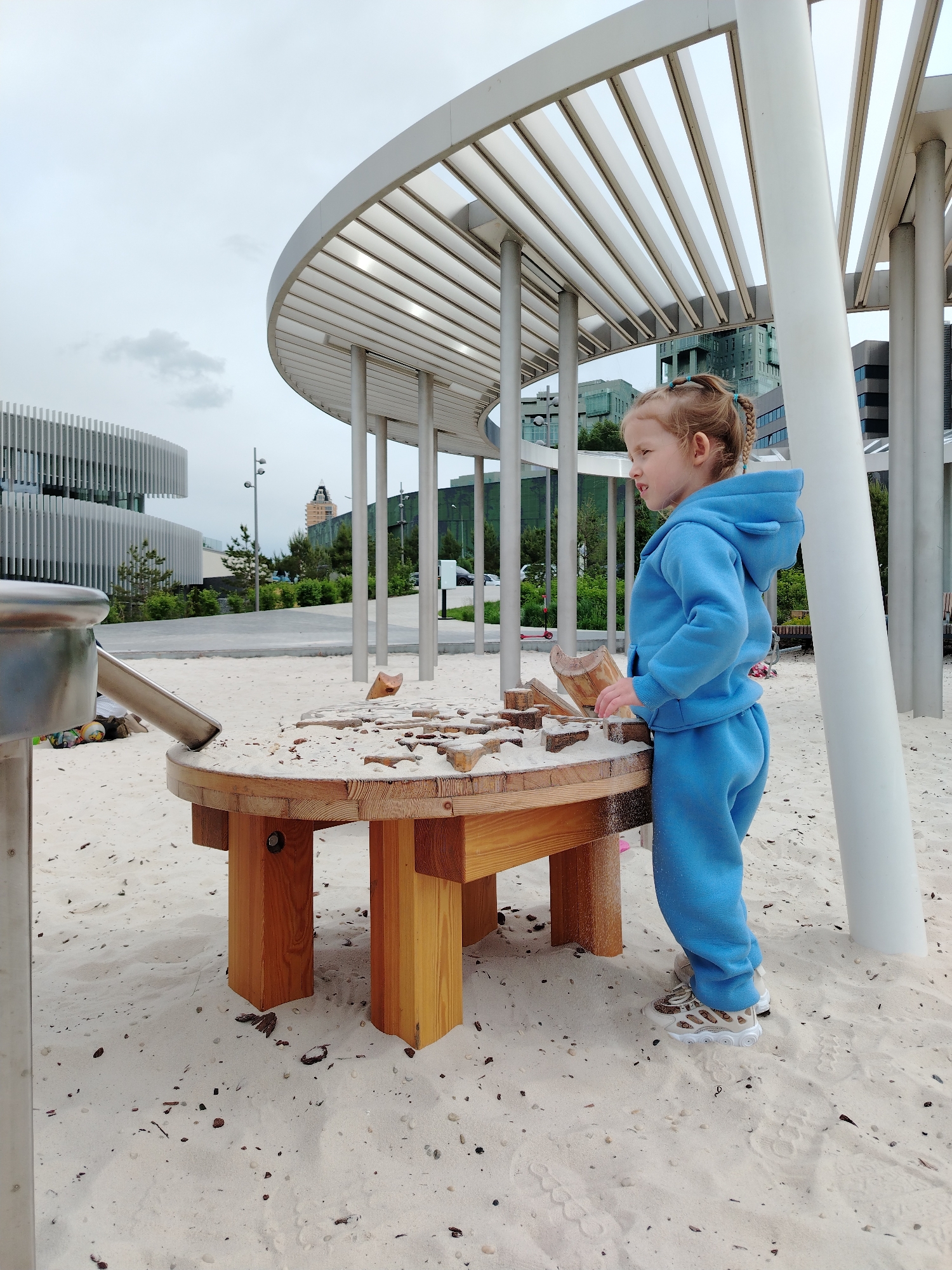 A young child in a blue hooded tracksuit plays at a wooden table partially filled with sand in a sandy park area in Kazan, Russia. The playground area is under a white metal structure supported by white columns. The child is blonde with hair styled in braids and is wearing patterned sneakers. In the background are modern buildings and greenery. The scene appears to be an outdoor play area within an urban environment.