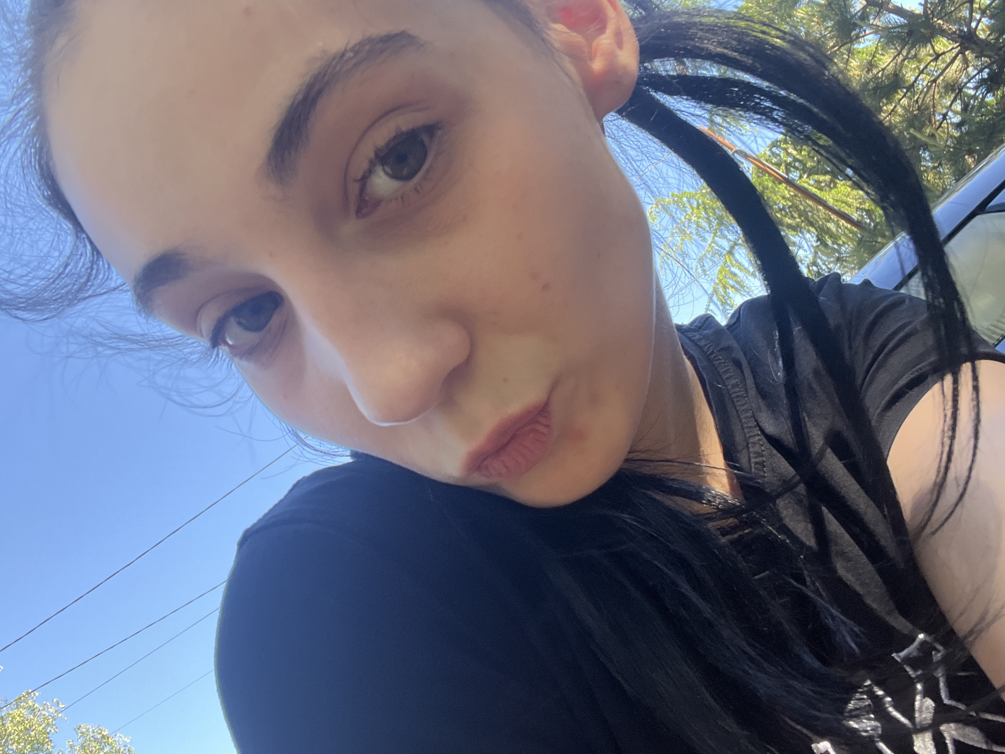 A close-up selfie shows a young woman with fair skin, dark hair in two pigtails, and dark eyebrows. She is wearing a black shirt with a graphic design. The background consists of a clear blue sky, some power lines, greenery, and a portion of a dark-colored vehicle. The location is likely outdoors. The city of Starozagorski bani, Bulgaria is not evident in this picture.