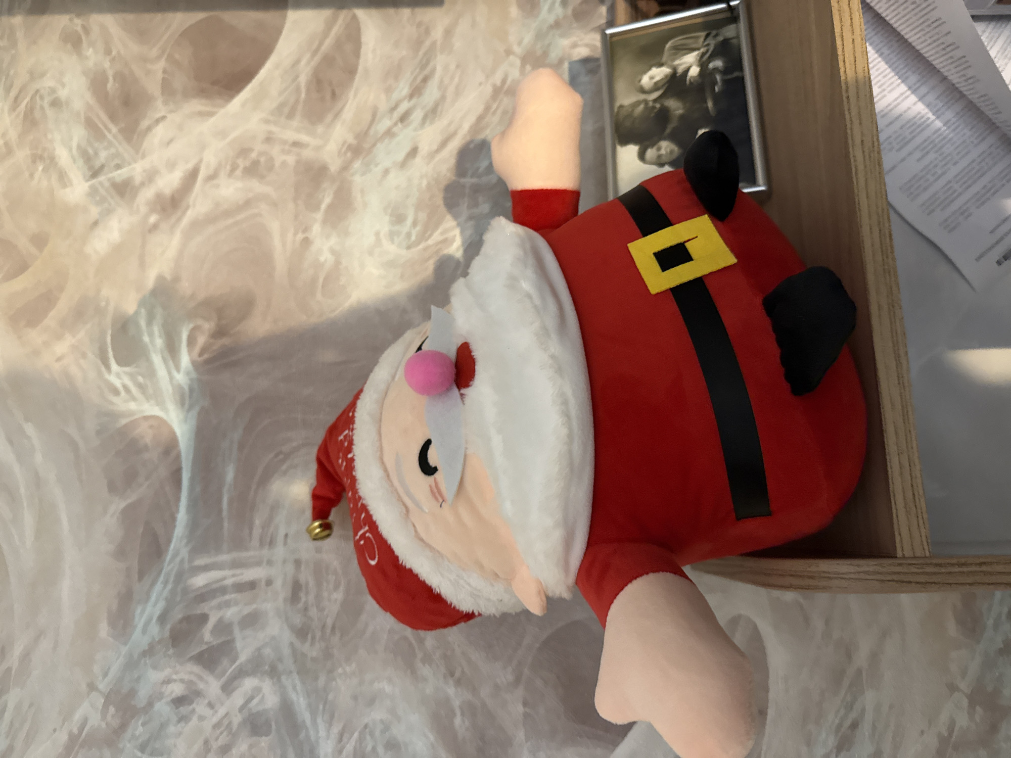 A stuffed Santa Claus toy is positioned on a wooden shelf. The toy is red and white with a black belt and yellow buckle. Behind the toy, a framed photograph shows three people. A patterned wallpaper or wall covering is visible in the background. There is a stack of papers on a lower shelf. The image does not provide any information about the location in the city of Saint Petersburg, Russia.