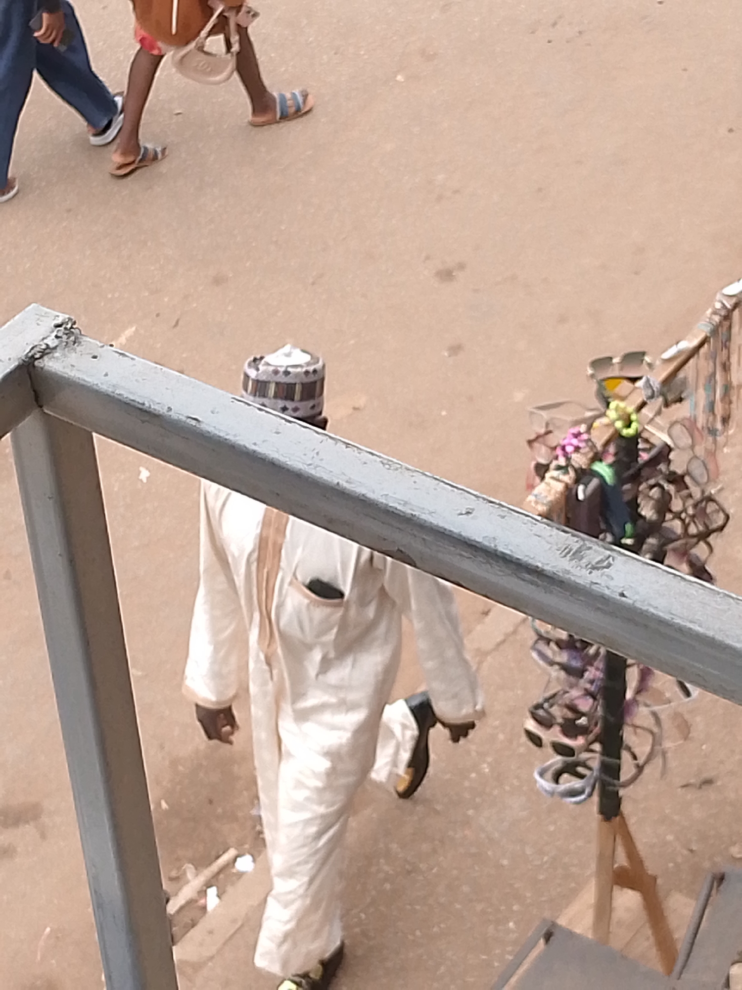 The image shows a view from an elevated position in Bukuru, Nigeria, looking down on a street scene.
People:
* A man in a white kaftan and a patterned kufi cap is walking away from the viewpoint. He has a dark complexion.
* Partially visible in the upper left corner are legs of two other people. One is wearing jeans and sneakers. The other is wearing sandals.
Objects:
* A grey metal railing frames the lower left side of the image.
* A display stand holding sunglasses and possibly other items is to the right.
* The street surface is light brown and appears to be paved.
Actions:
* The man in the kaftan is walking.
Environment:
* The scene is outdoors in daylight. The setting is a street in Bukuru, Nigeria.