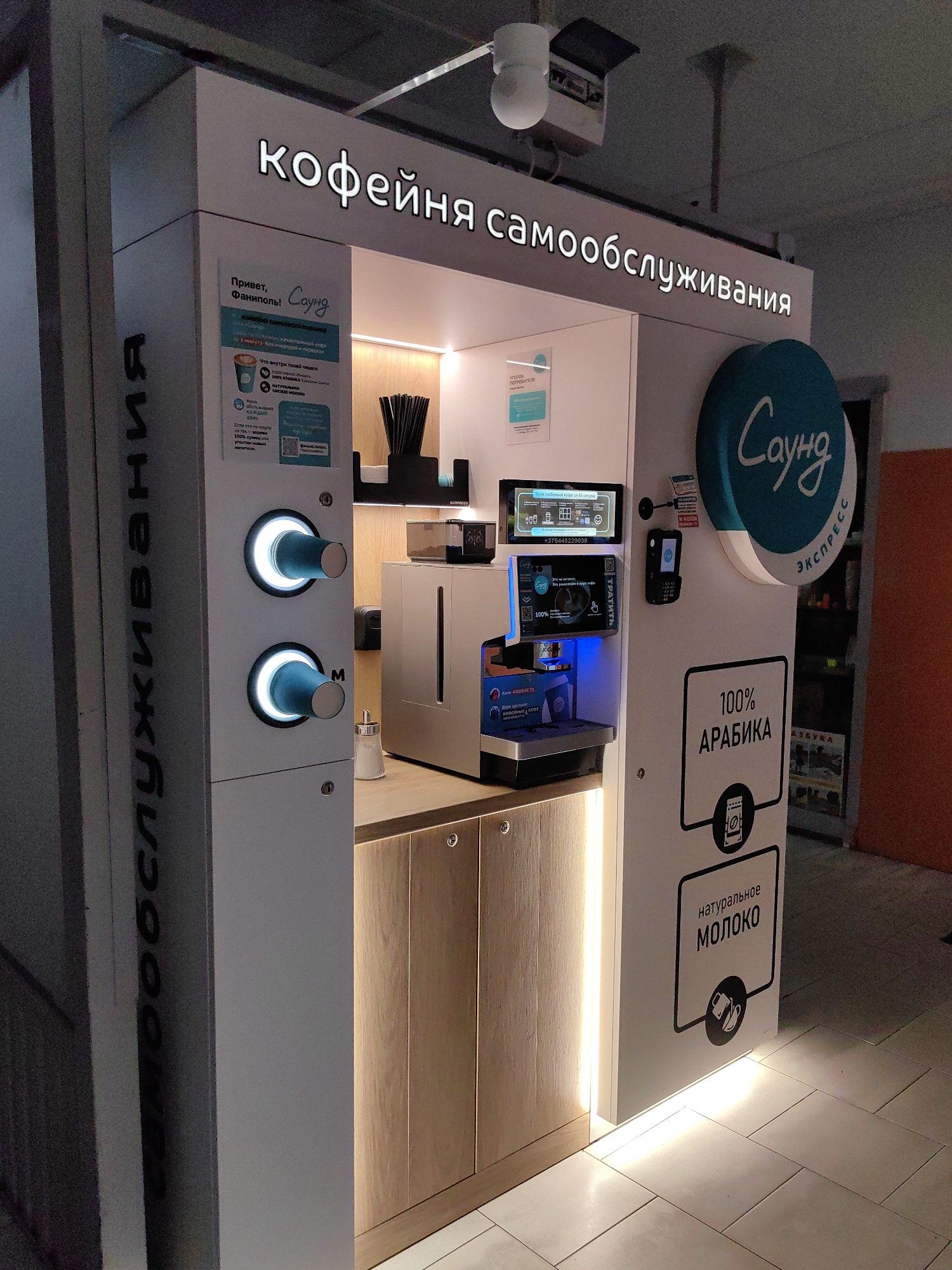 The image shows a self-service coffee kiosk in Fanipal, Belarus. The kiosk is white with illuminated signage. The main sign at the top reads "кофейня самообслуживания" (self-service coffee shop). To the left, there are two cup dispensers that are illuminated from within. Below them, text is vertically oriented and reads "ООО СПУЛИВАНИ" which appears to be a partial name or slogan. On the right side, a circular logo reads "Саунд ЭКСПРЕСС" (Sound Express). Below this, there are two framed panels: the top one has text that translates to "100% ARABICA" with an icon of coffee beans, and the bottom one reads "NATURAL MILK" with an icon of a milk jug.
In the center of the kiosk, there is a coffee machine with a touch screen interface above it, displaying various coffee options and a phone number. There are also straws and napkins on a shelf above the coffee machine. A payment terminal is mounted on the right side of the machine. The floor is tiled with light-colored square tiles. The overall lighting is warm and inviting, with illuminated strips along the bottom of the kiosk. The environment appears to be indoors, possibly in a public space like a mall or train station. There are no people visible in the image.