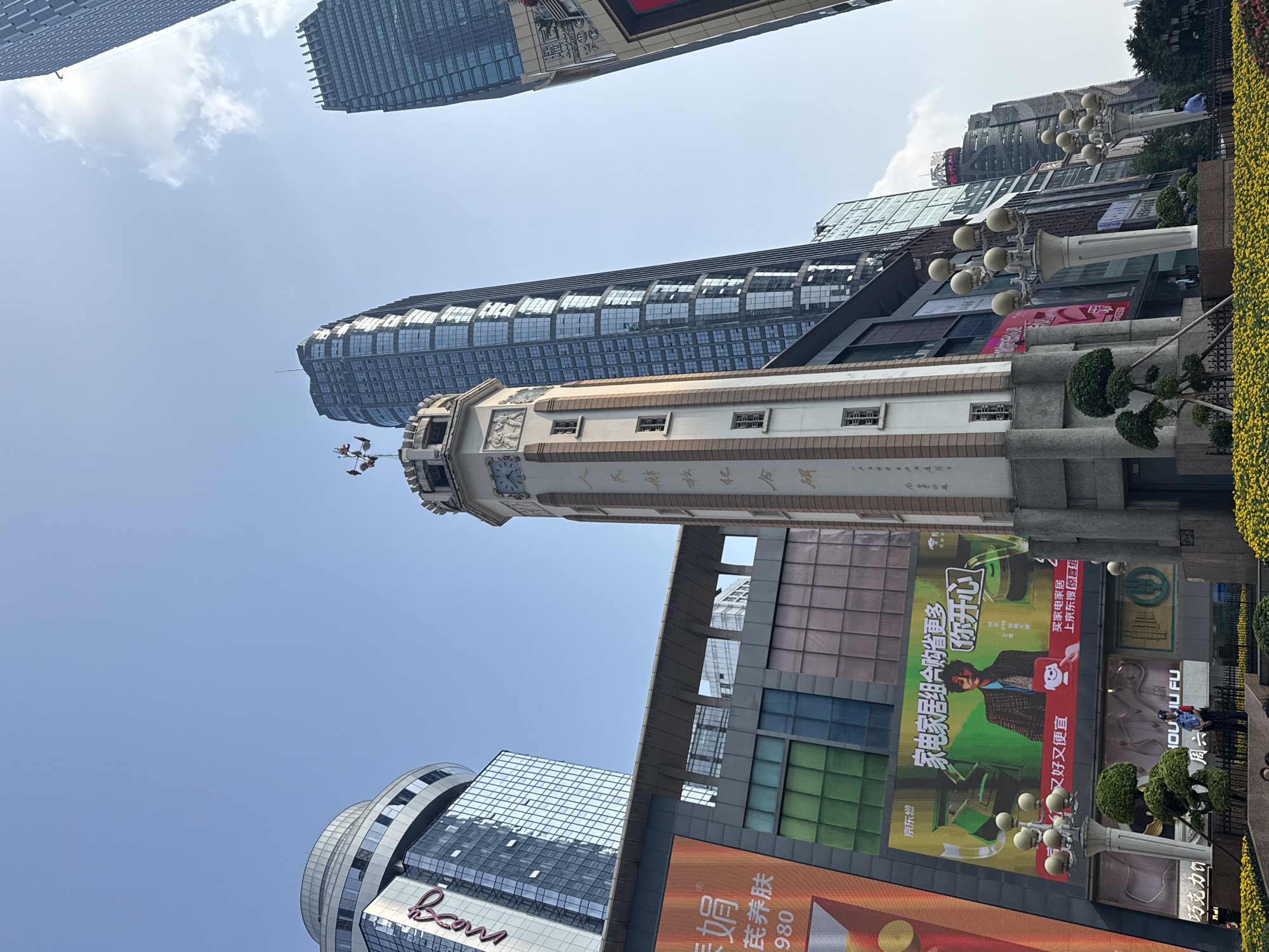 An outdoor, upward-looking shot captures a vibrant urban street scene, likely in Yuzhong District, China, under a bright, clear blue sky with sparse clouds. The image is dominated by the contrasting architecture of modern skyscrapers and a distinctive, ornate older tower, which appears to be a clock tower with what looks like a commemorative inscription in Chinese characters along its side, reading in part "...人民解放纪念碑..." (People's Liberation Monument). On its pinnacle, two figures are visible.
The modern buildings feature reflective glass facades, some with undulating designs, reflecting the sky. Street-level activity is hinted at by small figures and extensive commercial elements. Prominent in the foreground are several large digital billboards displaying advertisements. Visible Chinese text includes "Boom" on a building, and various promotions for "京东" (Jingdong), an e-commerce platform. One large billboard advertises "家电家居组合购省更多 你开心 又好又便宜" (Buy home appliances and furniture in a combination to save more, are you happy, good and cheap), along with phrases like "买家电家居 上京东搜" (Buy home appliances and furniture, search on Jingdong). Other visible text fragments include "娟", "芪养肤", "980", "518", "京东10大便", "HOUJU FU", "巧克力饮" (Chocolate drink), and "周六" (Saturday).
The scene is well-lit, indicating daytime. Modern streetlights with multiple globes and manicured shrubbery add to the urban aesthetic. The low angle of the shot emphasizes the towering height of the structures against the expansive sky.