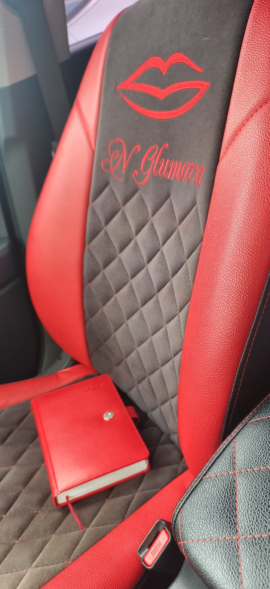 The image captures a close-up view of a car's interior, specifically focusing on a highly customized front seat. The seat features a striking two-tone design, primarily in red and a dark grey or black, suggestive of a personal and vibrant aesthetic.
The central panel of the seat is upholstered in a dark grey, suede-like material with a diamond-quilted pattern, highlighted by red stitching that traces the grid. Embroidered prominently on this central panel are bright red lips, an artistic graphic, positioned above a name or word in red cursive script that appears to read "N Glimora" or "N Glumora." The side bolsters and accents of the seat are made from a rich, textured red leather, providing a strong contrast and sense of luxury. A black textured cushion or armrest with matching red stitching is visible on the far right.
Resting on the seat's dark grey panel, near the bottom left, is a vibrant red notebook or planner. It has a metallic button clasp and a red ribbon bookmark protruding from its spine. Faintly embossed on the cover, the year "2026" is visible, suggesting it's a future-dated item or an annual planner.
A seat belt buckle is partially visible in the bottom right, with a red "PRESS" button set into a black frame. The scene is brightly lit, indicating daytime, and appears to be stationary. The location context for this image is Фоки, Russia. No people are visible in the frame, and no specific activity is taking place beyond the presentation of these car interior details and objects.