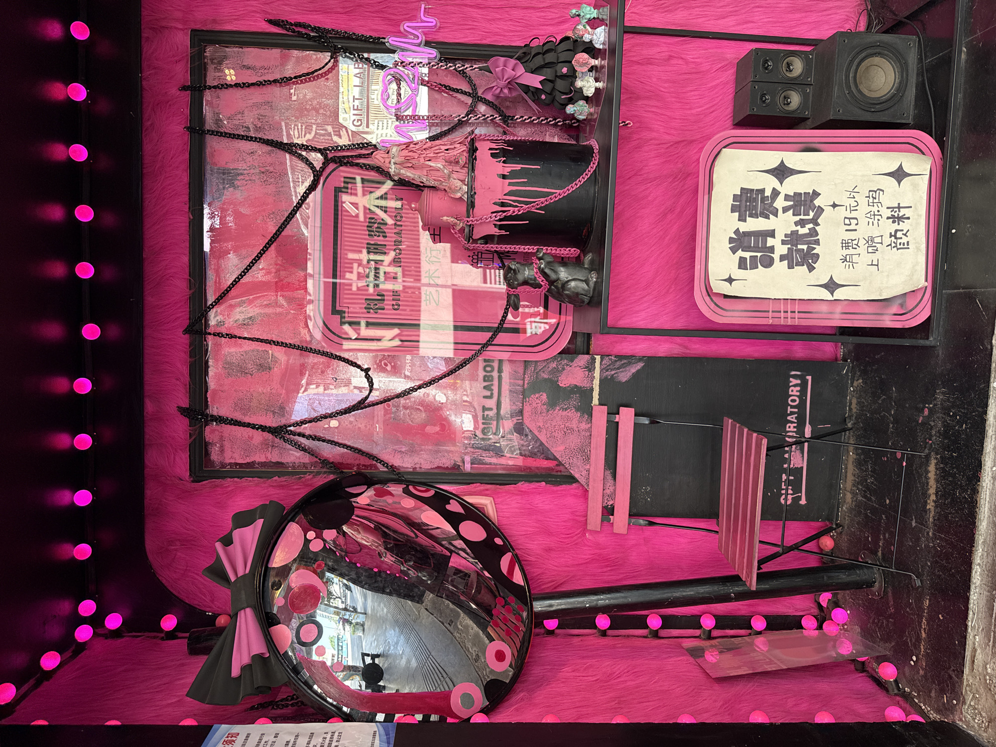 A photograph of a gift display in the Jiulongpo District of Chongqing, China, showcases a whimsical assortment of items in a vivid pink setting.
The backdrop consists of a bright pink faux fur material. Several objects are displayed: a framed piece with a pink abstract design, adorned with black chains; a black container with pink paint seemingly dripping down; and a round mirror with pink and black polka dots and a bow. There's also a small black speaker and a sign with Chinese characters on it. A miniature pink chair sits on one side. The scene is illuminated by bright pink spherical lights strung around the perimeter of the display.