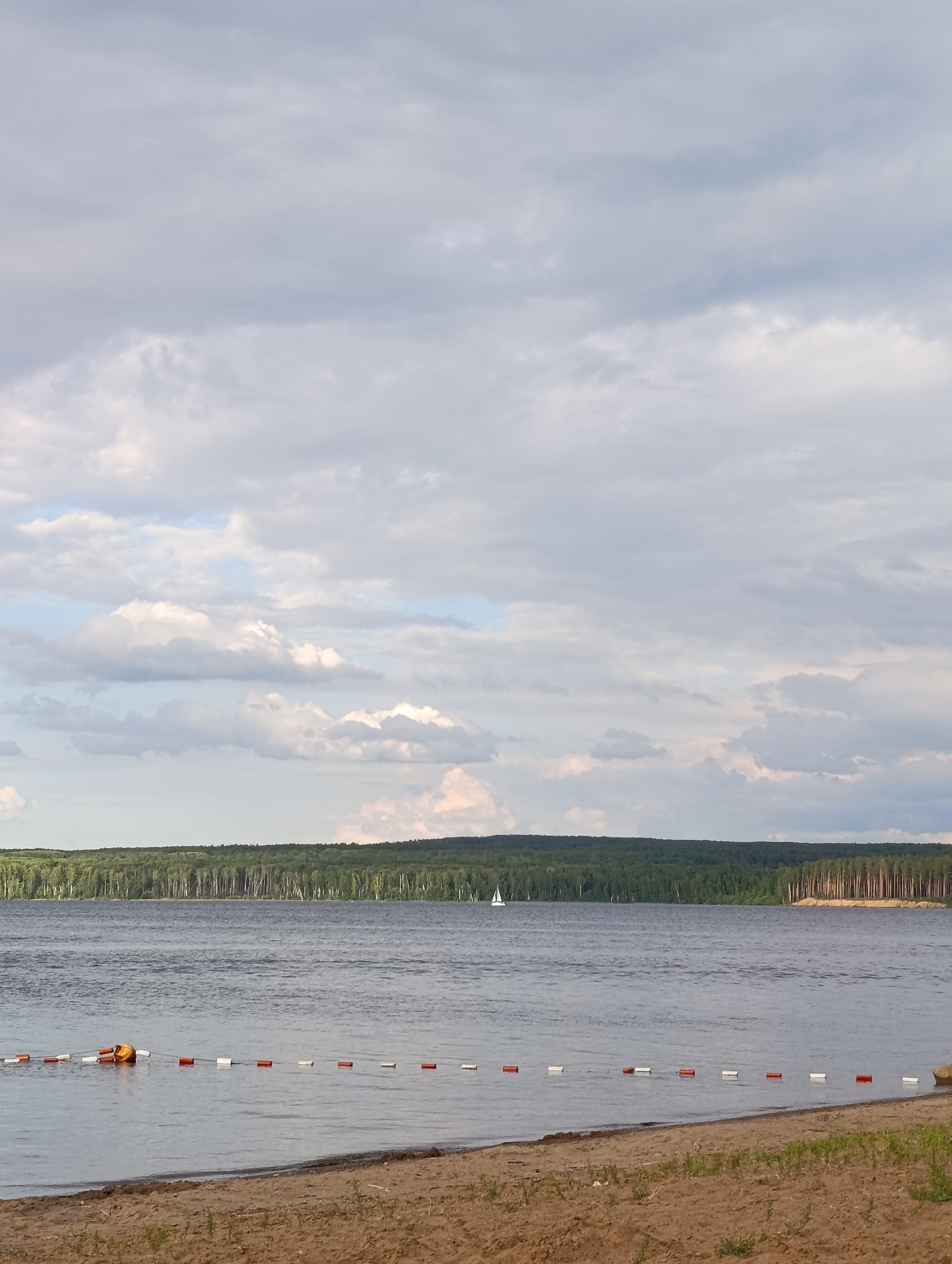 A wide shot captures a serene lake landscape. The foreground features a sandy beach with sparse green vegetation. The calm water stretches out, marked by a line of buoys with alternating red and white floats, and a larger orange and yellow buoy. In the distance, a dense, dark green forest lines the far bank of the lake. A white sailboat is visible on the water, and the sky above is filled with a mix of grey and white clouds, suggesting an overcast day. The overall impression is one of natural tranquility.