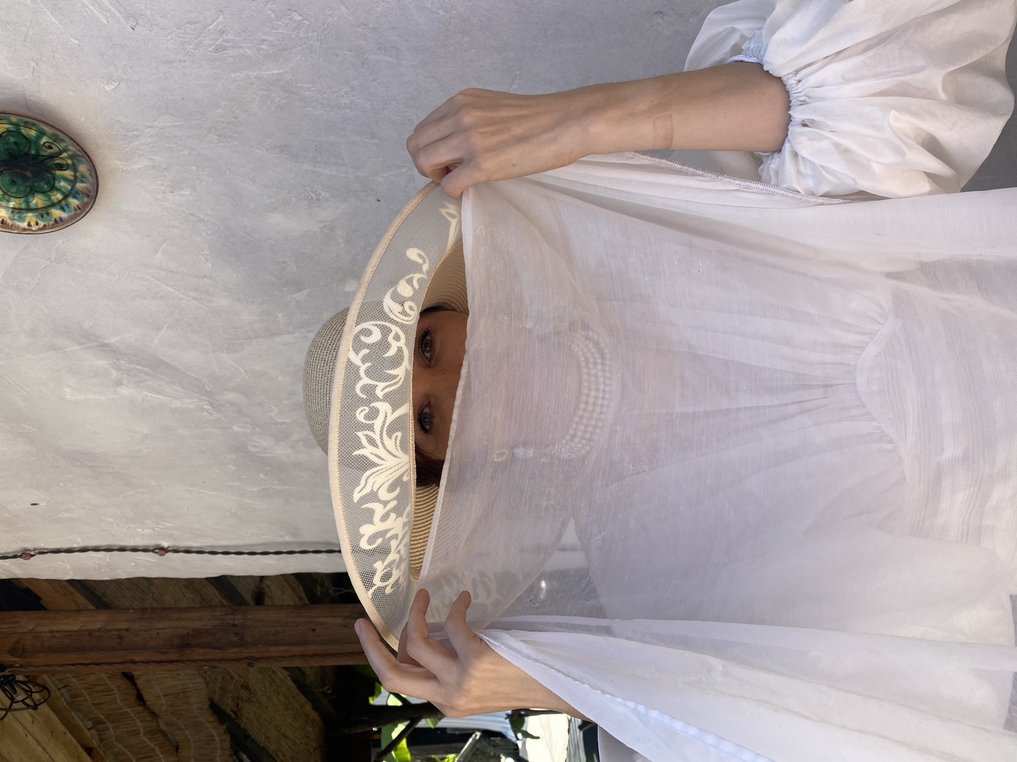 The image shows a person wearing a large, straw hat and a sheer white veil that covers their face and chest. The veil is draped over their head and shoulders, obscuring most of their features except for their eyes, which are visible through the fabric. The person is wearing a white garment with voluminous sleeves, and a string of pearls is visible beneath the veil. The background consists of a textured, off-white wall and a wooden beam supporting a thatched roof. A decorative, oval-shaped object with green and yellow patterns is affixed to the wall. The scene appears to be outdoors, possibly in a covered patio or veranda. The lighting suggests a bright, sunny day. The individual's hands are visible, one on each side, holding the veil in place. The overall impression is one of mystery or playful concealment. There are no clear indications of the specific city, but the architectural style and vegetation might suggest a warm climate.