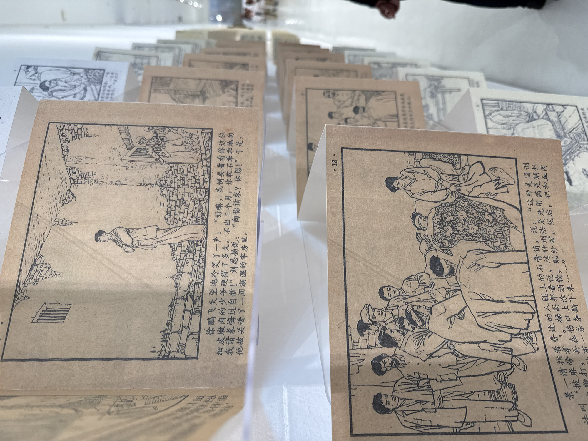 The image shows a collection of small, paper-covered blocks, each featuring black and white line drawings and text in Chinese. The subjects of the drawings appear to be historical or fictional narratives, featuring people in scenes. The text provides context or captions for the images. The setting is an indoor exhibit, likely in an art gallery or museum. The precise location cannot be determined beyond the possible location of Chongqing High-tech Zone, China. The objects are the blocks themselves, arranged in rows on a light-colored surface. The action is viewing or display, suggesting an exhibit or presentation.