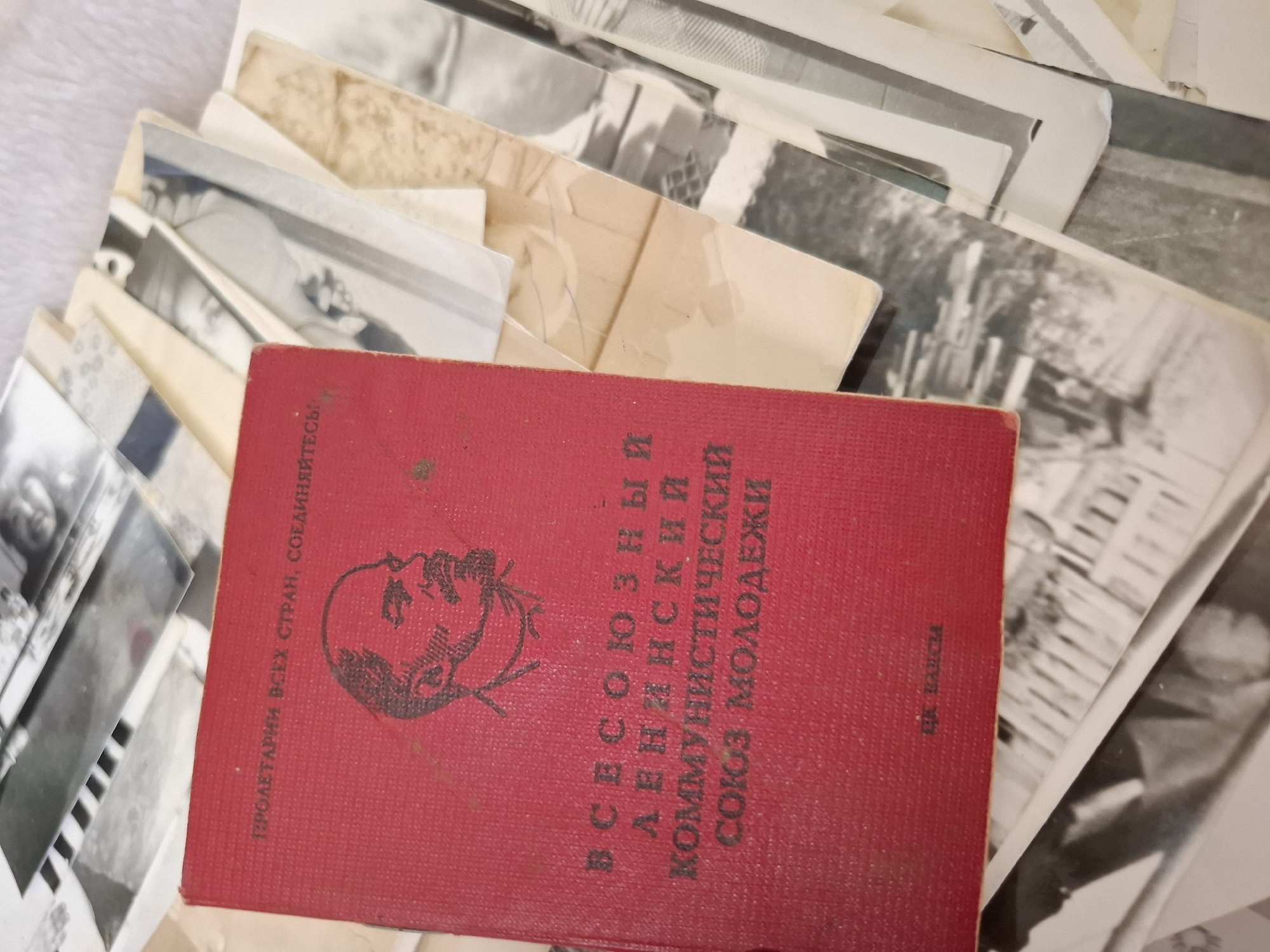 The image shows a red book with Cyrillic text and an image of Vladimir Lenin on its cover, seemingly a Soviet-era youth organization membership card (Komsomol). The book is surrounded by a stack of photographs, which are mostly black and white. In the lower-right corner of the book, the text reads "И.Е. Бахси". The text on the book reads "Пролетарии всех стран, соединяйтесь!" and "ВСЕСОЮЗНЫЙ ЛЕНИНСКИЙ КОММУНИСТИЧЕСКИЙ СОЮЗ МОЛОДЕЖИ". The context suggests the setting is somewhere in a Soviet-era setting. The city of Амзя, Russia, is not directly discernible from the image.