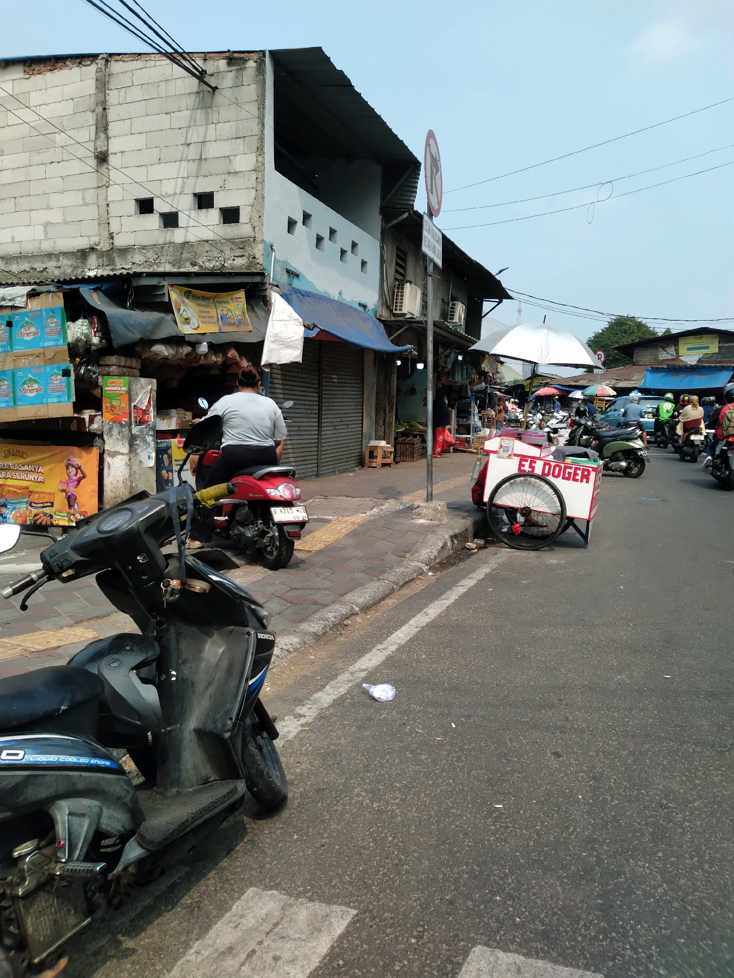 The image captures a street scene in South Tangerang, Indonesia, likely a bustling local market area. In the foreground, a black Honda scooter is parked on the asphalt road. Further down the street, a woman is seated on a red scooter. Adjacent to the sidewalk, a white and red food cart, labeled "ES DOGER," is positioned. Several other scooters and motorbikes are visible on the road, suggesting ongoing traffic. Buildings with visible signage and awnings line the street, indicating a commercial or residential area. The sky is clear and blue, with overhead power lines crossing the frame. A white umbrella is open, providing shade over part of the street. The overall impression is a snapshot of daily life in a busy Indonesian urban environment.
