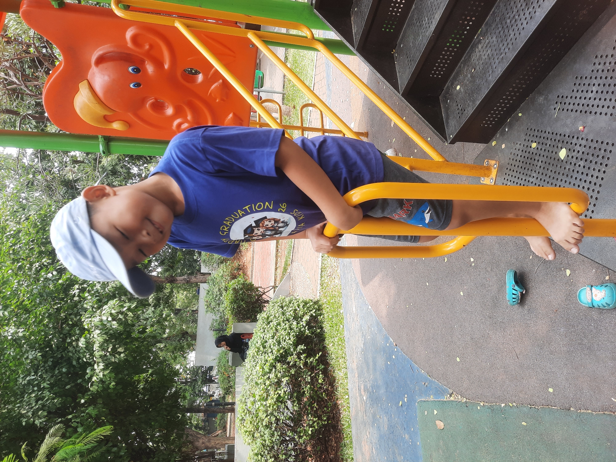 In a public park in North Jakarta, Indonesia, a young boy wearing a blue baseball cap and a blue t-shirt with a graduation-themed graphic is standing on a playground structure. He is gripping a yellow metal bar, with his head tilted down and a slight smile on his face. He is wearing gray shorts with a shark graphic. Black metal stairs are visible to his right. On the grey rubberized ground in front of him are a blue toy car and two blue children's sandals. In the background, lush green trees and bushes surround the playground. Further back, a woman in a black hijab is seated on a bench, with other people and park amenities visible in the distance.