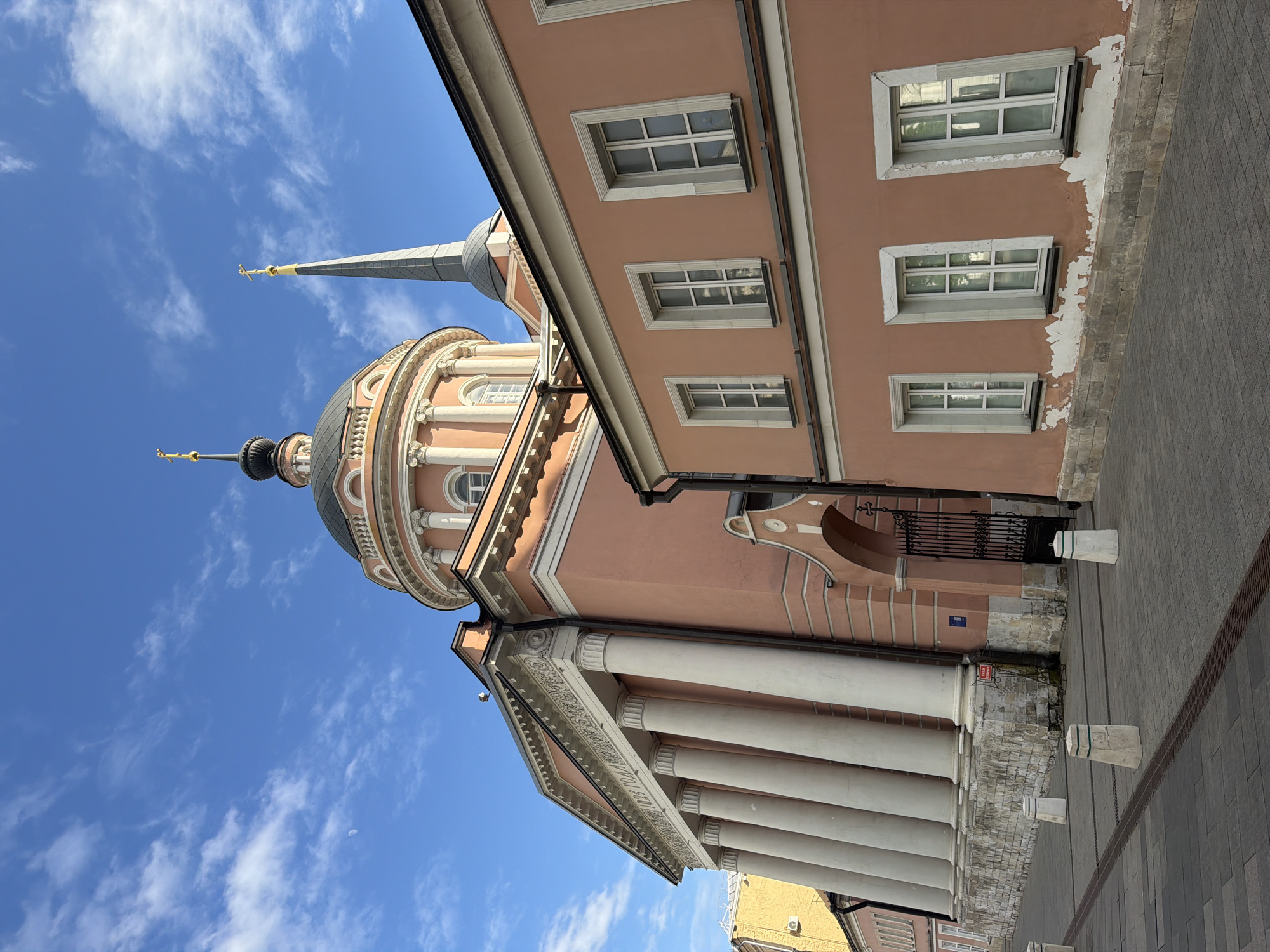 The image depicts the Church of St. Nicholas in Kotelniki, Moscow, Russia, as seen from a low angle. The church features a pink facade, white columns, and a dome topped with a spire. A building with pink walls and white-framed windows is adjacent to the church. The sky is blue with scattered white clouds. The ground is paved with grey stones.