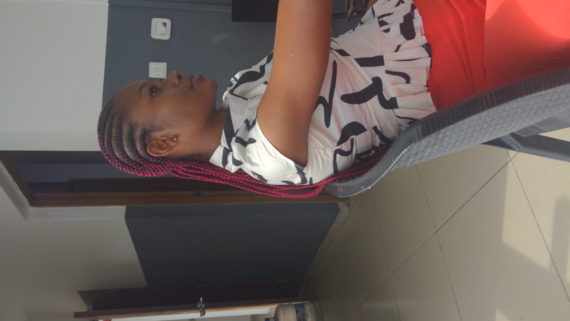 An indoor, side-profile shot captures a young woman seated in a dark grey, woven-textured chair, looking upwards and slightly to her right. The woman has dark skin and her hair is styled in intricate cornrows, dyed a distinct reddish-purple or maroon color. A small gold stud earring is visible in her right ear.
She is wearing a white short-sleeved top with bold, abstract black patterns, which appears to be ruffled or pleated at the front. Her lower garment is a bright orange or red skirt or trousers. The woman's expression is calm and attentive, with her lips slightly parted.
The background features an interior wall painted in two distinct sections: a darker grey or charcoal color on the upper portion, and a lighter off-white or light grey on the lower section. Two white electrical outlets or switches are mounted on the darker wall. The floor is covered with light-colored, possibly cream or beige, tiles with visible grout lines. A wooden frame or trim is visible along the base of some walls. A partially obscured large plastic water jug with a blue lid is seen in the lower right corner. The lighting suggests it is daytime. This scene depicts a casual indoor setting, likely in Abuja, Nigeria, possibly a waiting area, office, or home. No text is clearly legible.