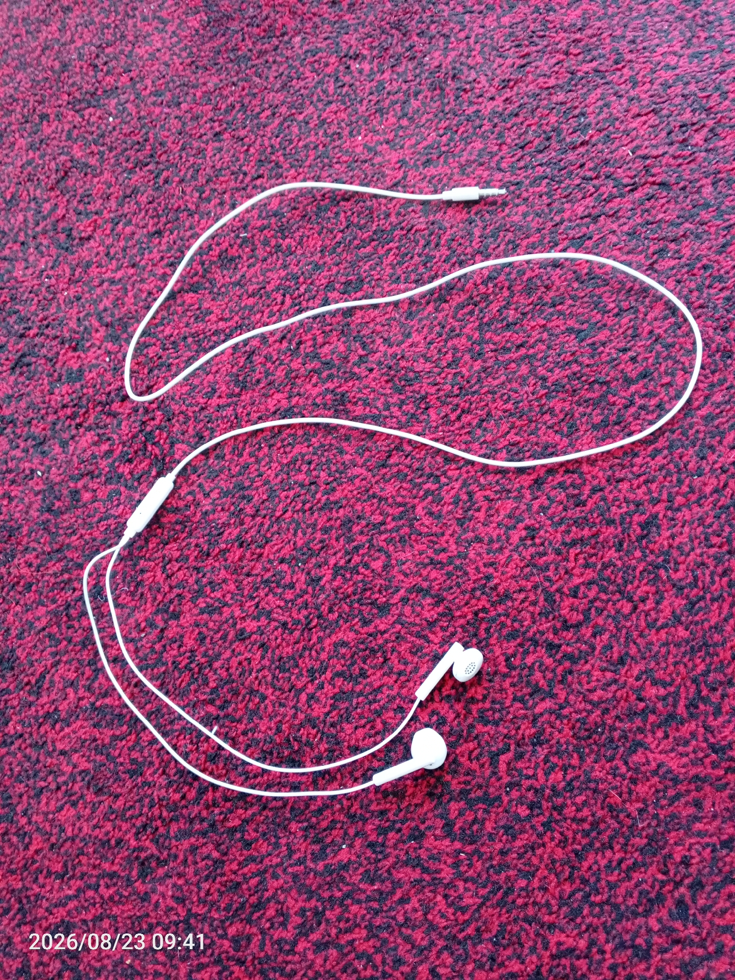 The image shows a pair of white wired earbuds with a microphone on a red and black speckled carpet. The earbuds are placed in the lower right quadrant of the image, with the cord extending diagonally upwards and to the left. The jack of the earbuds is located in the upper left quadrant of the image. The carpet appears to be a typical indoor rug. The timestamp "2026/08/23 09:41" is visible in the bottom left corner of the image. There are no people or any visible indication of the location being in Yokari-Kushtamgali, Uzbekistan.