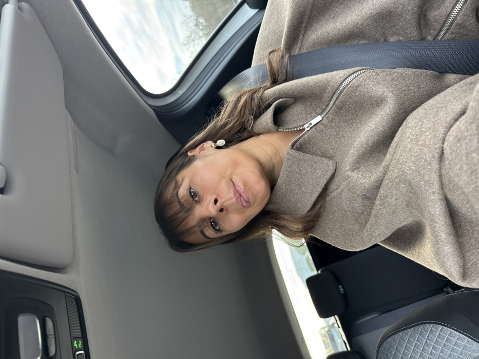 A woman is inside a car, angled towards the camera. She has brown hair with bangs and is wearing a brown zippered jacket with a seatbelt across her chest. Her facial expression appears playful, with her tongue slightly sticking out. A small, light-colored earring is visible. The car's interior is predominantly gray, with a glimpse of the car window and a sky visible in the background.