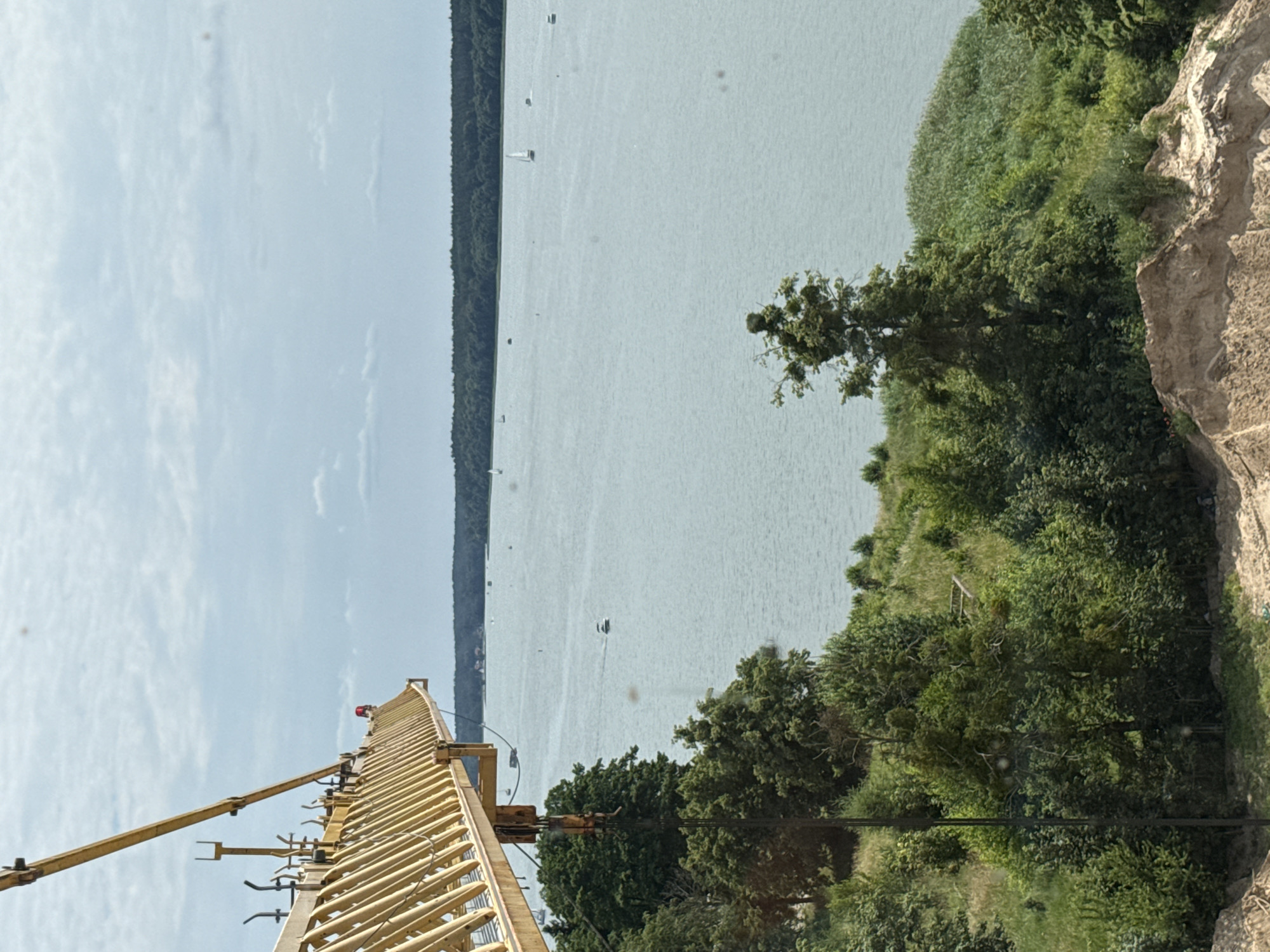 This is a landscape photograph taken outdoors on a bright, cloudy day. The image is oriented vertically. In the foreground, a yellow construction crane extends from the lower left corner upwards and towards the right. The crane appears to be assembled and ready for use, with its lattice structure clearly visible. Above the crane, the sky is a pale blue with scattered white clouds.
The middle ground of the image features a wide body of water, possibly a lake or a bay. Several small white sailboats are dotted across the surface of the water, suggesting a recreational or sporting activity. The water is calm and reflects the light from the sky. Along the far edge of the water is a dense, dark green line of trees, forming a natural barrier.
The right side of the image shows a steep, rocky embankment covered with lush green vegetation and trees. The textures of the rocks are rough and varied, with some areas appearing lighter and others darker. The trees are dense and leafy, creating a vibrant green contrast with the pale water and sky.
The overall impression is one of a serene natural setting with a touch of human industry. The bright sky and calm water suggest pleasant weather, likely during daylight hours. The presence of sailboats hints at activities like sailing or boating. The rocky embankment and dense foliage indicate a natural landscape, possibly a lakeside or coastal area. No text is visible in the image.
