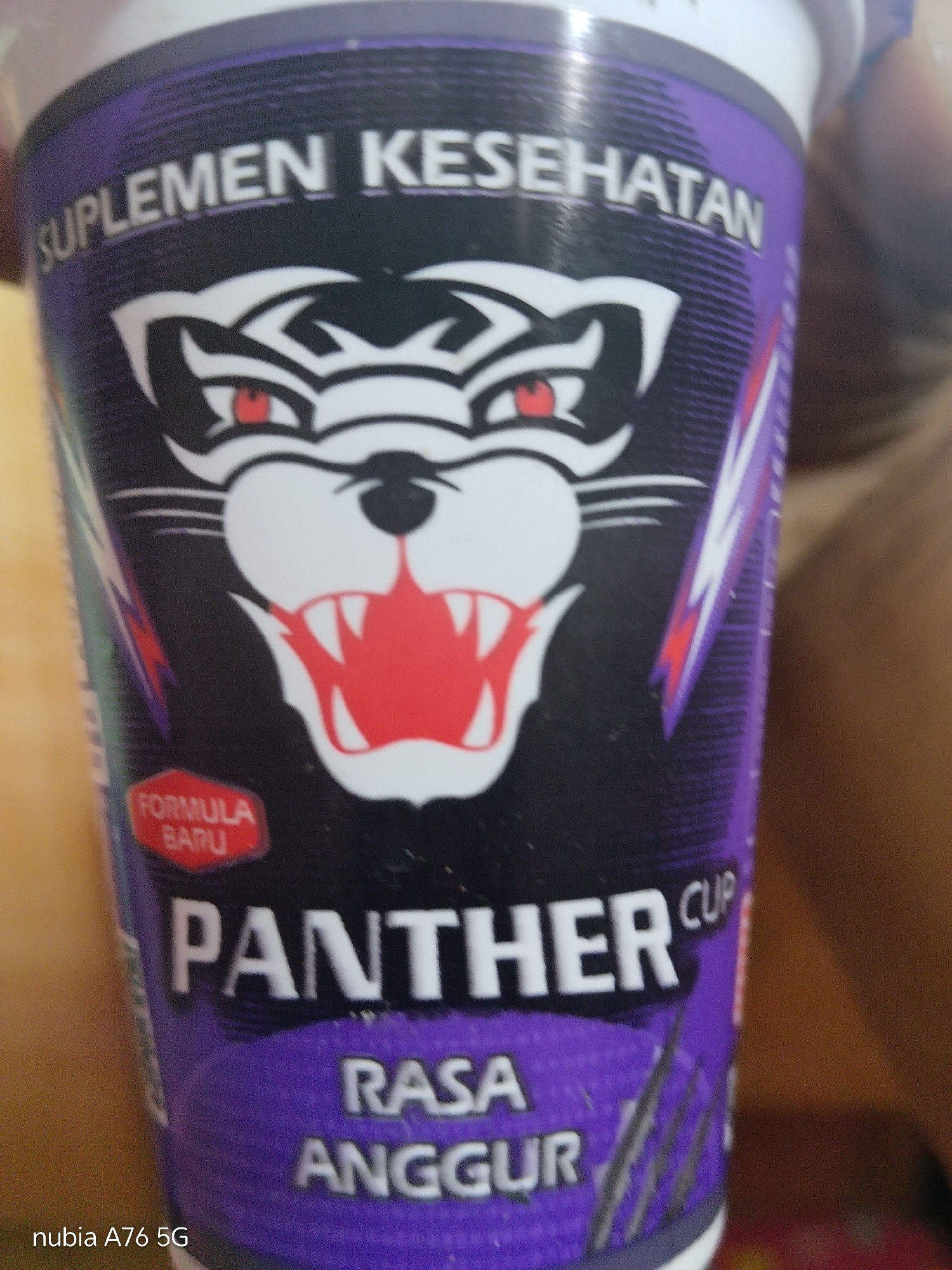 The image shows a close-up of a purple and black cup labeled "SUPLEMEN KESEHATAN FORMULA BARU PANTHER CUP RASA ANGGUR". The cup features a stylized black panther's face with red eyes and an open mouth revealing red fangs. The background is blurred and appears to be an indoor setting with a wooden surface visible. The text "nubia A76 5G" is present in the bottom left corner, indicating the device used to capture the image. There is no information or visual cues in the image that would allow for determining its location in Cakung, Indonesia.
