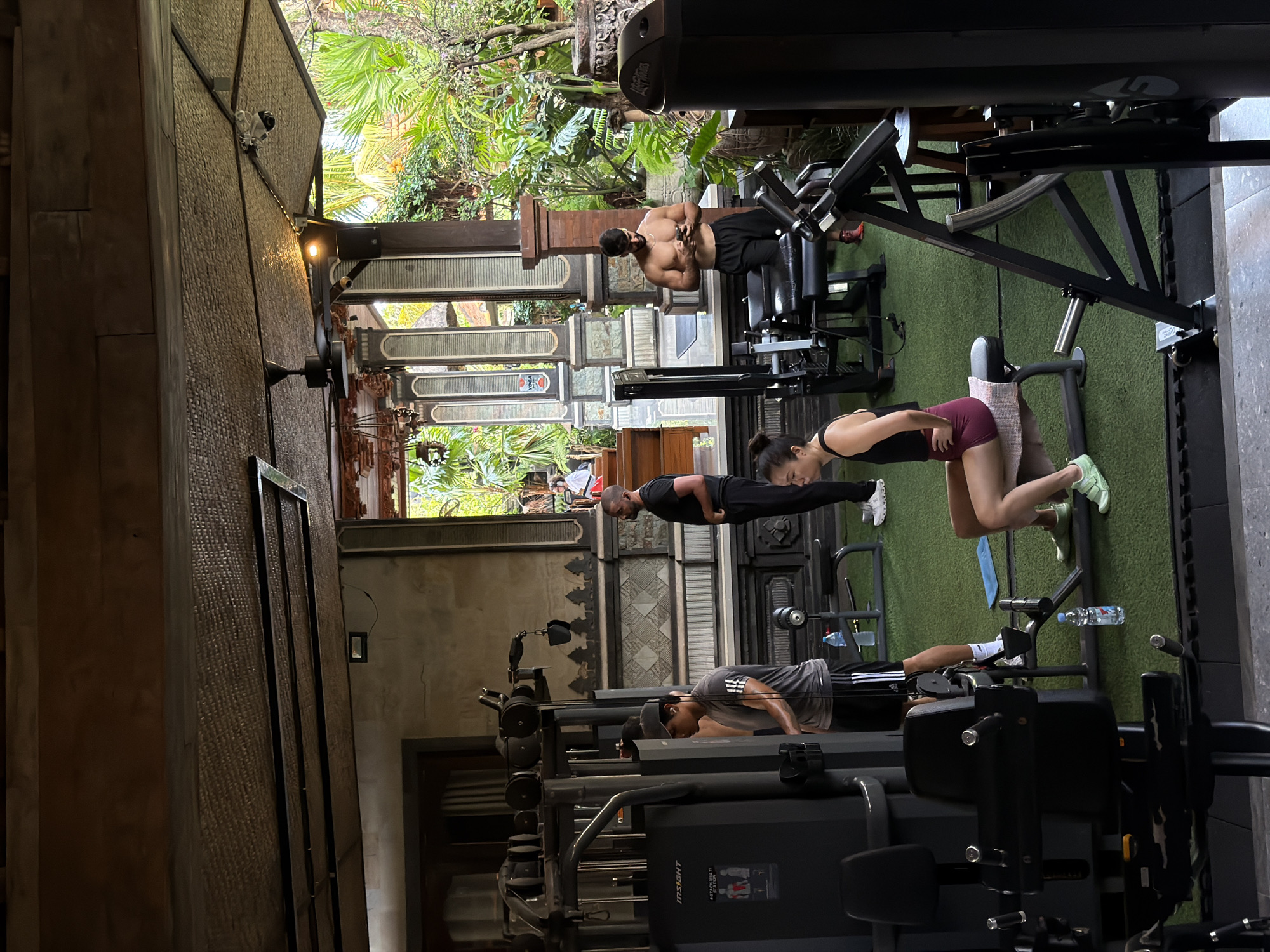 An open-air gym in North Kuta, Indonesia, is captured during daytime, with natural light illuminating the interior and lush tropical foliage visible outside. The gym features a mix of modern fitness equipment and traditional architectural elements, including carved wooden columns and patterned wall panels. The floor transitions between green artificial turf and a darker surface.
At least five individuals are exercising. In the foreground to the right, a woman in a black top and dark red shorts performs an exercise on a leg machine, wearing light green sneakers. Near the center, a man in a black t-shirt and black pants stands upright, possibly resting between sets. To the left, another man in a grey t-shirt, dark shorts, and a baseball cap is seen performing push-ups. Further back, a shirtless, muscular man in black shorts stands near gym equipment. A fifth person's head and shoulder are partially visible near the standing man in black.
Visible gym equipment includes multi-stack weight machines, one of which has "INSIGHT" and "4 STACK MULTI GYM" printed on it, and a dumbbell rack. Water bottles are scattered on the floor. A security camera is mounted on a wall. A sign displaying "YOGA" is partially visible in the background. The scene suggests a focused workout environment within a resort-like or traditional setting.
