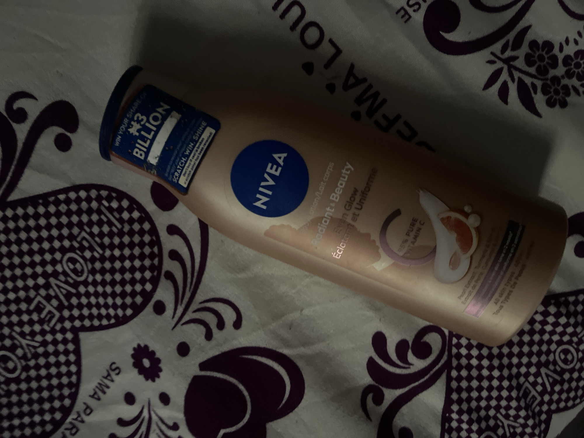 A Nivea Radiant + Beauty Even Glow (Éclatant et Uniforme) body lotion bottle, light peach in color with a blue cap, is positioned on a patterned fabric surface. The bottle's label displays "95% PURE VITAMIN C" and mentions Pearl Extract, Turmeric, and Sheer. A promotional sticker on the cap reads "WIN YOUR SHARE OF N3 BILLION" with the Naira symbol, along with "SCRATCH, WIN, SHINE," and an advisory about broken seals. The fabric background is white with dark purple/maroon repeating patterns, including checkered geometric shapes, stylized floral motifs, and partial text such as "I LOVE YOU," "SAMA PARA," and "DERMA LOU." The scene is illuminated by indirect light, casting soft shadows. No individuals or specific actions are depicted. The promotional sticker indicates the product is marketed in Nigeria. The image lacks specific visual identifiers for the city of Abuja.
