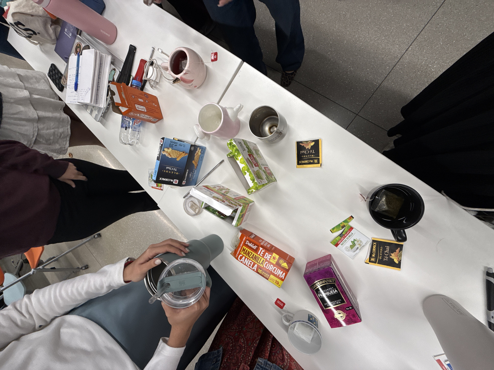 An overhead shot captures a white table laden with various items, primarily focused on tea and beverages, suggesting a casual break or gathering in Mexico City, Mexico. Several people are partially visible around the table. One person, wearing a white long-sleeve shirt, holds a gray Stanley tumbler. Another person in dark clothing is seated, and a third person's legs in blue jeans and leopard print shoes are visible.
The table is cluttered with multiple tea boxes and ingredients for hot drinks. Notable objects include a pink Owala water bottle, a small pink pig-shaped mug, a silver travel mug with "MATION" inscribed, and several tea boxes. These include "MCCORMICK BLENDS Té Chai" (some showing "20 sobres" and "40g"), "NATURE'S HEART. MATCHA VAINILLA", an orange box of "Doblett TÉ DE MANZANILLA CÚRCUMA CANELA" with "16 sobres 19.2g", and a purple box of "DAVENPORT GOURMET FRAMBUESA CONTENT. 40 g". Other items include an orange box of "Splenda" sweetener packets, a plastic water bottle, a white notebook with a blue pen, and charger cables. A black mug with a tea bag steeping is also visible. The setting appears to be an indoor space with a gray tiled floor.
