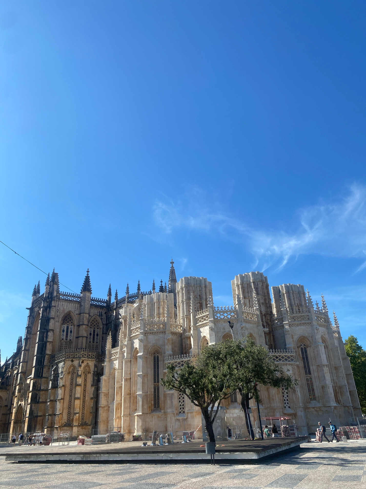 The image shows the Batalha Monastery in Batalha, Portugal. The monastery is a UNESCO World Heritage site, characterized by its Gothic and Manueline architectural styles. The left side of the structure exhibits darker stone, suggesting age and possibly different construction periods, with pointed arches and intricate window tracery. The right side of the monastery is lighter in color and features elaborate buttresses, pinnacles, and decorative stonework, including large, multi-story towers with crenellated tops.
In the foreground, a paved plaza area with a patterned surface extends towards the monastery. A raised wooden platform is situated in the plaza, with a trash receptacle and a lone tree nearby. Several people are visible walking in the plaza, some appearing to be tourists. Construction materials, including scaffolding and stacked metal structures, are present in front of parts of the monastery, indicating ongoing maintenance or restoration work. The sky is a clear, vibrant blue with wispy white clouds, suggesting a sunny day.