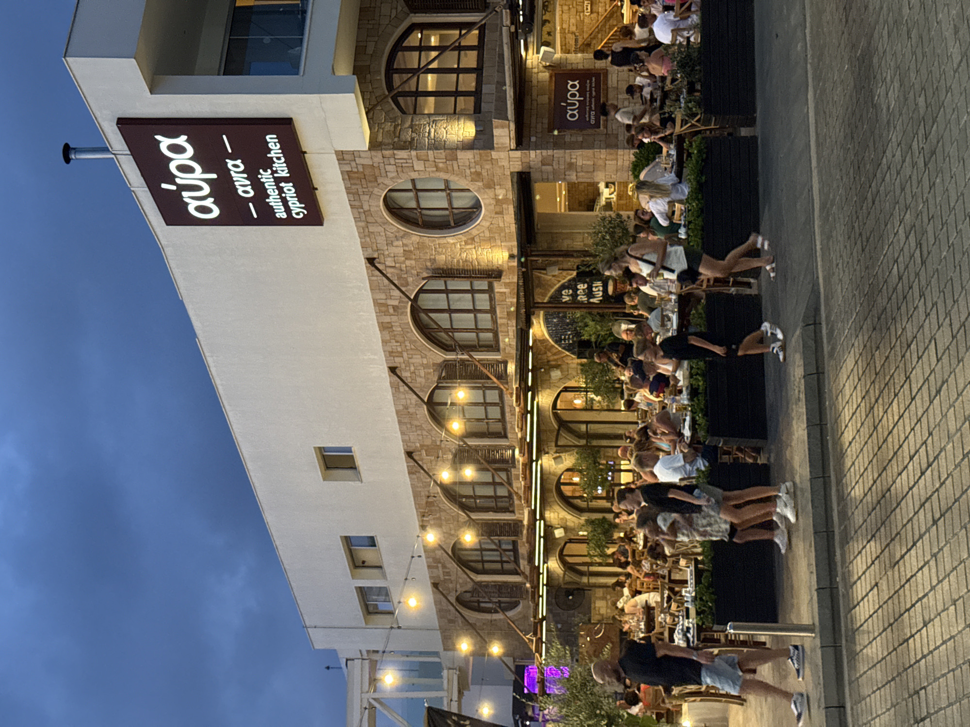 The image depicts the exterior of a restaurant named "avra - authentic cypriot kitchen" located in Ayia Napa, Cyprus. The restaurant has a stone facade with arched windows and is illuminated by string lights. A prominent sign with the restaurant's name is visible on the upper right corner. Several people are seated at tables outside the restaurant, engaged in dining and socializing. A few individuals are walking on the cobblestone sidewalk in front of the establishment. The sky is a deep blue, suggesting it is either dusk or early evening.