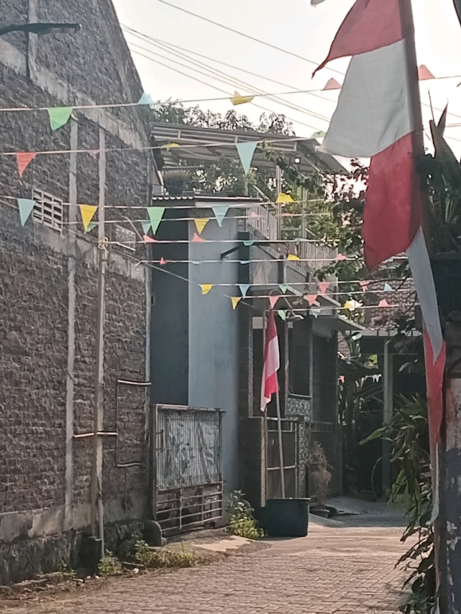The image depicts a narrow street lined with buildings in Semarang, Indonesia, adorned with small, colorful triangular flags strung across the scene. A large red and white flag, likely the Indonesian flag, is prominently displayed on the right side of the image, partially obscuring a building. The buildings themselves appear to be constructed from brick and have simple architectural styles, with some showing signs of wear. Vegetation, including trees and bushes, is visible in the background, suggesting a residential or mixed-use area. The street is paved with cobblestones, and the overall atmosphere suggests a local community celebrating an event. There are no people visible in the frame.