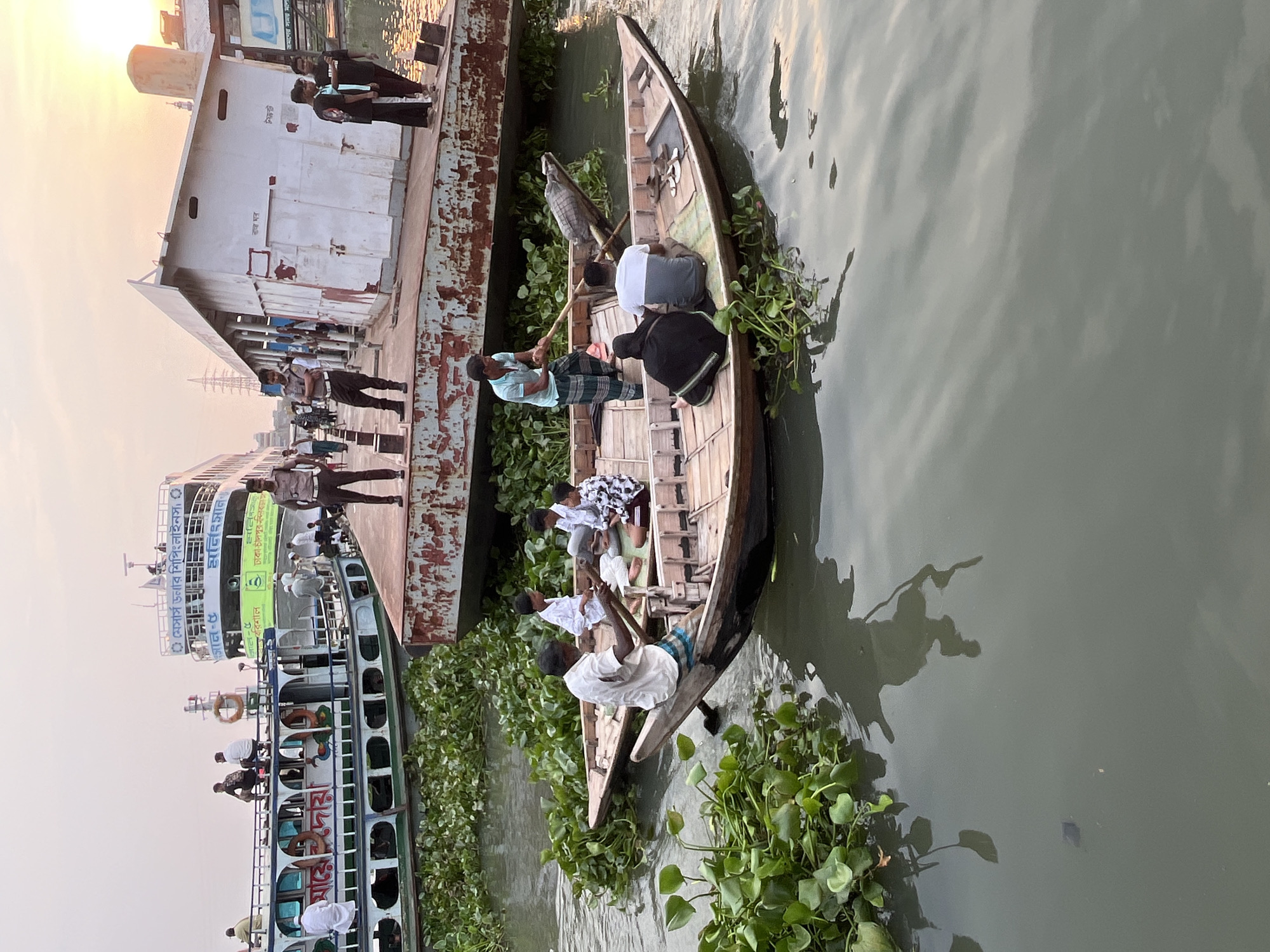 Here is a detailed description of the image:
**Location:** The scene is set in Dhaka, Bangladesh. This is indicated by the signage on the boats, which is written in Bengali script.
**Environment:** The image shows a waterway, likely a river or canal. There are boats alongside a pier. Water hyacinth is prevalent in the water.
**People:** There are several people in the image. Some are boarding or disembarking from a larger boat on the left. A smaller rowboat in the foreground contains several occupants.
**Subjects and Objects:**
* A larger boat is moored to the pier on the left side of the image.
* A small, narrow rowboat is visible in the foreground, occupied by at least five individuals. The boat is surrounded by water hyacinth.
* A building is situated on the pier, possibly a terminal or waiting area.
**Actions and Interactions:**
* People are boarding/disembarking the larger boat.
* The occupants of the rowboat appear to be navigating the waterway, with at least one person using an oar or pole.
* The location is a busy transportation hub, with boats moving people to and from different locations.
**Details:**
* The light suggests it may be near sunrise or sunset.
* The pier appears to be made of concrete and metal.
* The larger boat has multiple decks and is equipped with life rings.