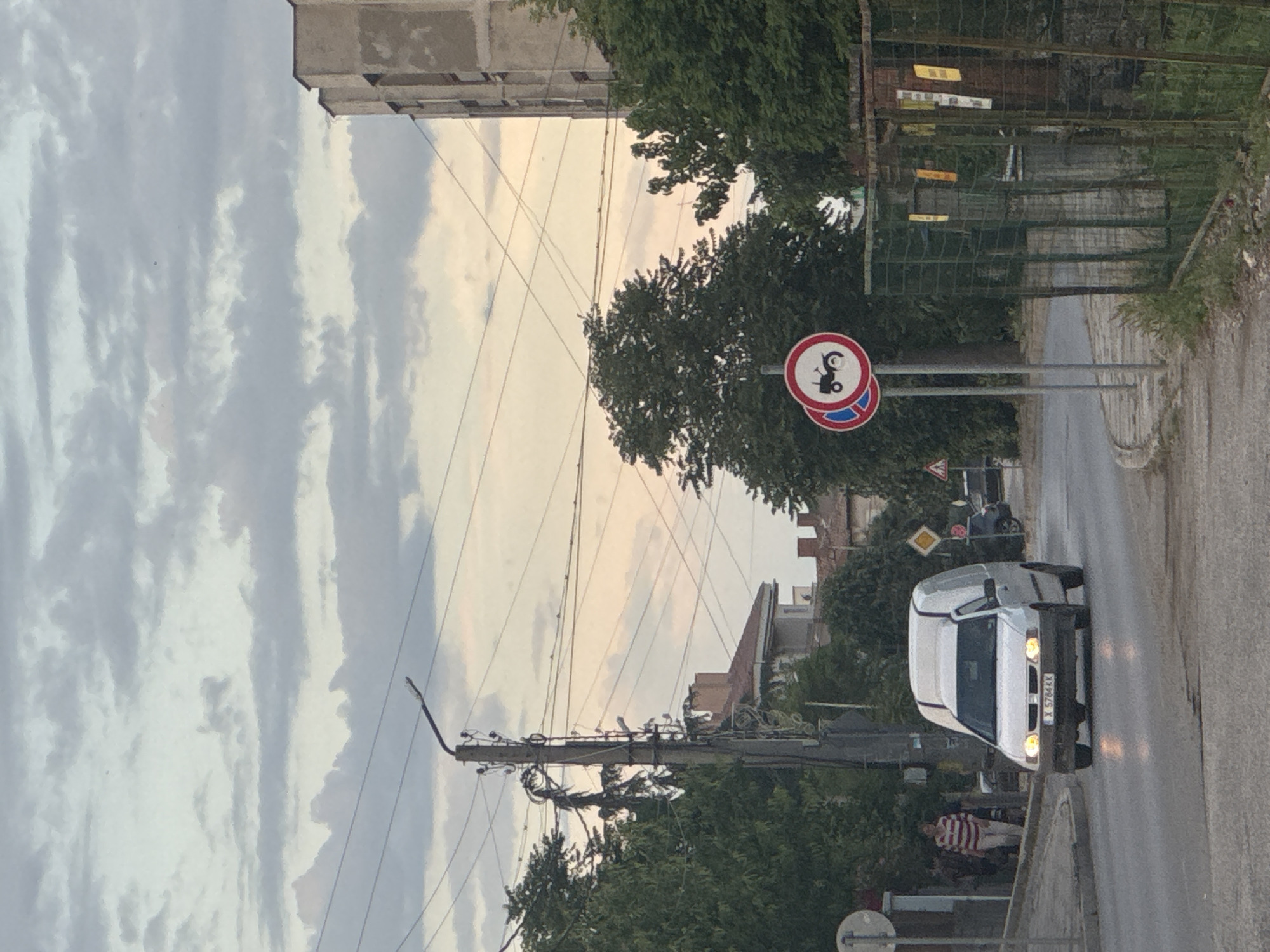 The image shows a street scene in Ivaylovgrad, Bulgaria. A white van is driving on a wet road. On the left side of the road, there is a sign prohibiting motorcycles and mopeds. An electrical pole with multiple wires is visible. In the background, there are buildings and trees. The sky is cloudy. A person in a red and white striped shirt is visible on the left side of the road. The van's license plate appears to read "X 5764 KK." A yield sign is visible near the motorcycle and moped prohibition sign.
