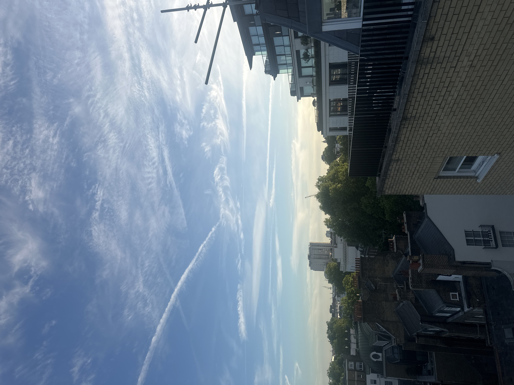 The image shows a daytime urban scene, likely in London, United Kingdom, given the architectural style. The sky is predominantly blue with scattered cirrus clouds and several contrails. From the vantage point of a high floor, a cityscape unfolds. Buildings of varying heights and architectural styles are visible, from modern white structures with balconies to older brick buildings with pitched roofs. A dense line of trees follows the street. A tall, modern building is visible in the distance. A television antenna sits on top of one of the buildings. The light suggests a sunny day, although the scene is slightly hazy. There are no people visible.
