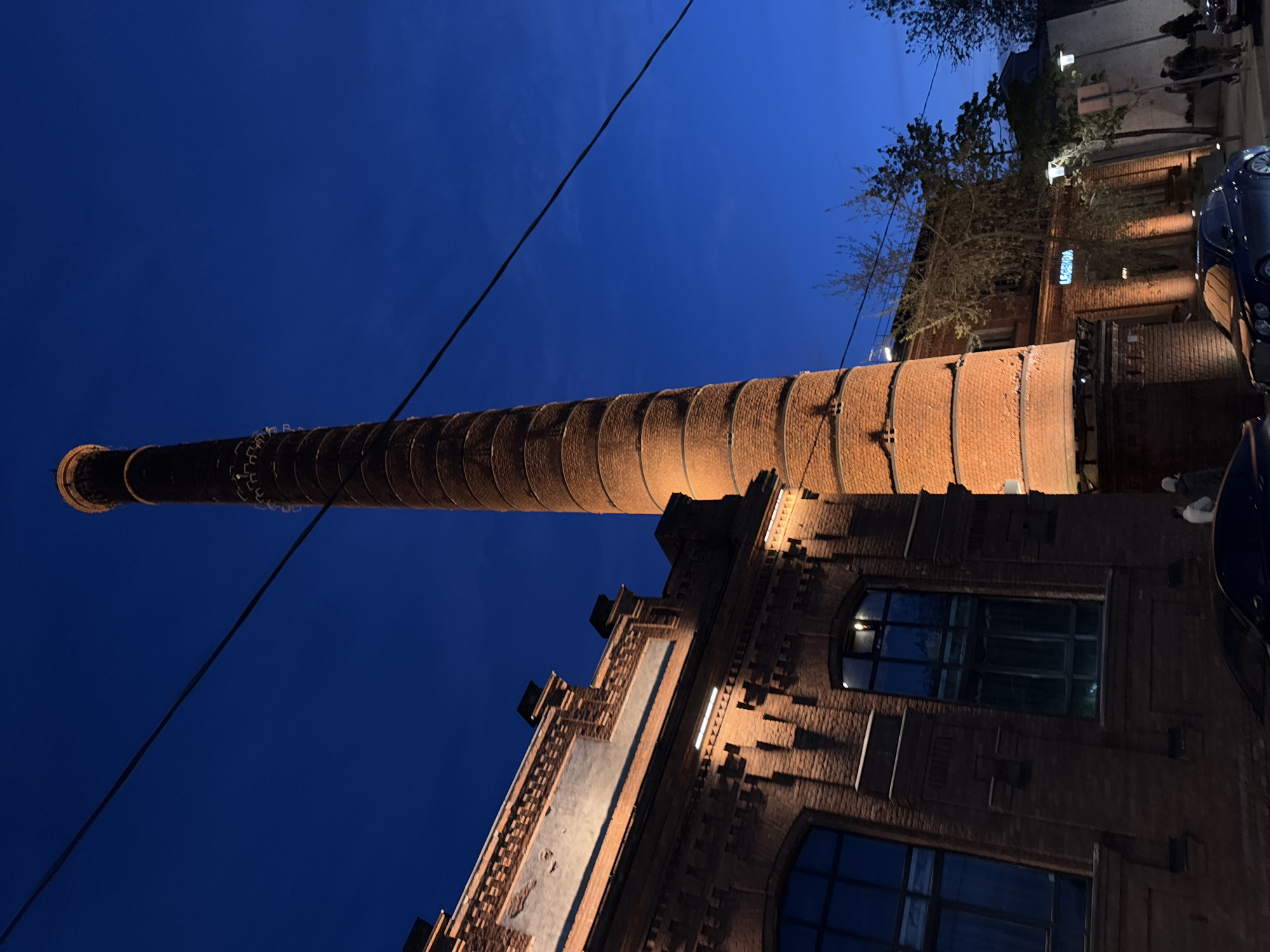 Here is a description of the image:
The image shows a tall, brick smokestack rising from a brick building against a dark blue sky. The building has multiple windows with arches and decorative brickwork. A powerline runs across the sky near the top of the stack. In the background, there are other buildings, trees, and a glimpse of people and a car. The photo appears to have been taken at night or during dusk.
This is most likely the Red Banner Factory in Saint Petersburg.